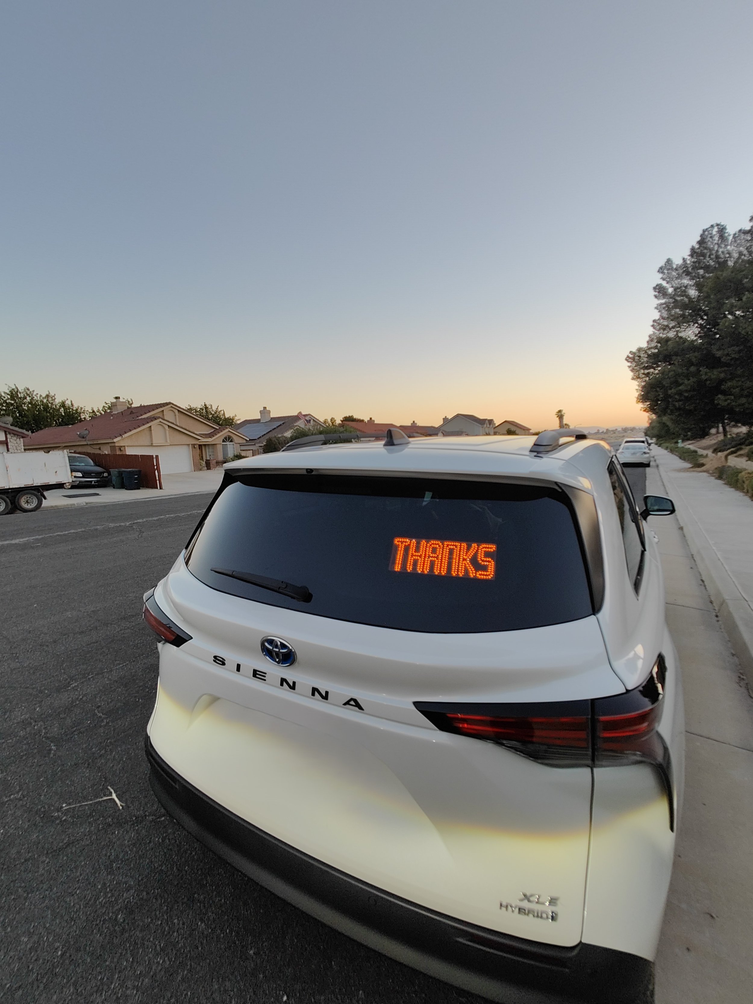 A white Toyota Sienna hybrid minivan parked on a suburban street at sunset, with an electronic sign on the rear window displaying the word 'THANKS' in orange LED lights.