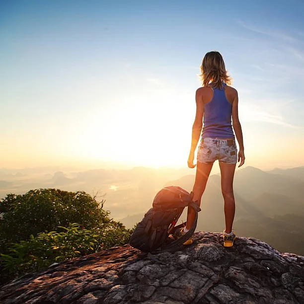 A woman standing on a mountain peak at sunrise, with a backpack beside her, overlooking a scenic mountain landscape.