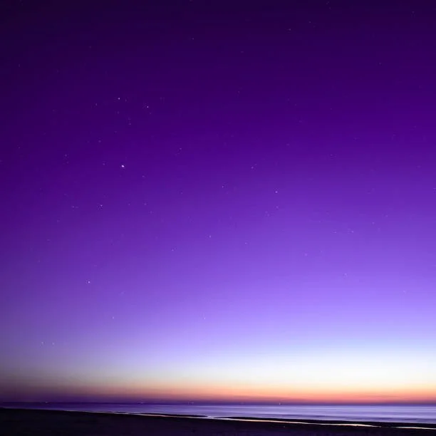 Night sky with stars over a calm body of water and a distant horizon with a gradient sunset or sunrise colors.