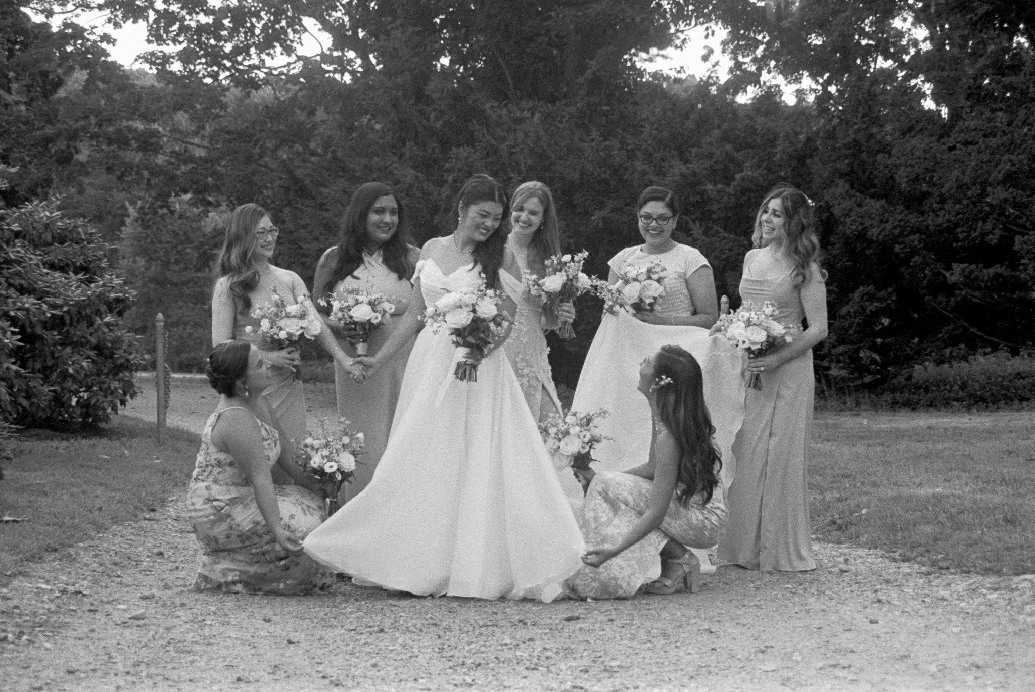 Six women in wedding dresses holding bouquets, surrounding a bride in a garden or park setting.