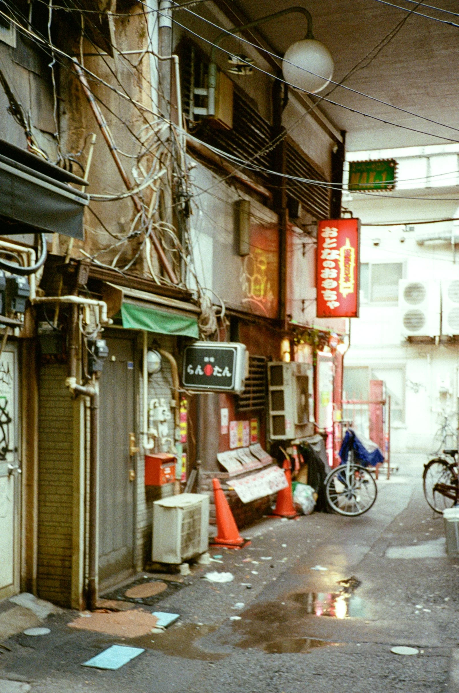 An alleyway in Japan with cluttered storefronts, tangled electrical wires, bicycles, garbage, and puddles on the ground.