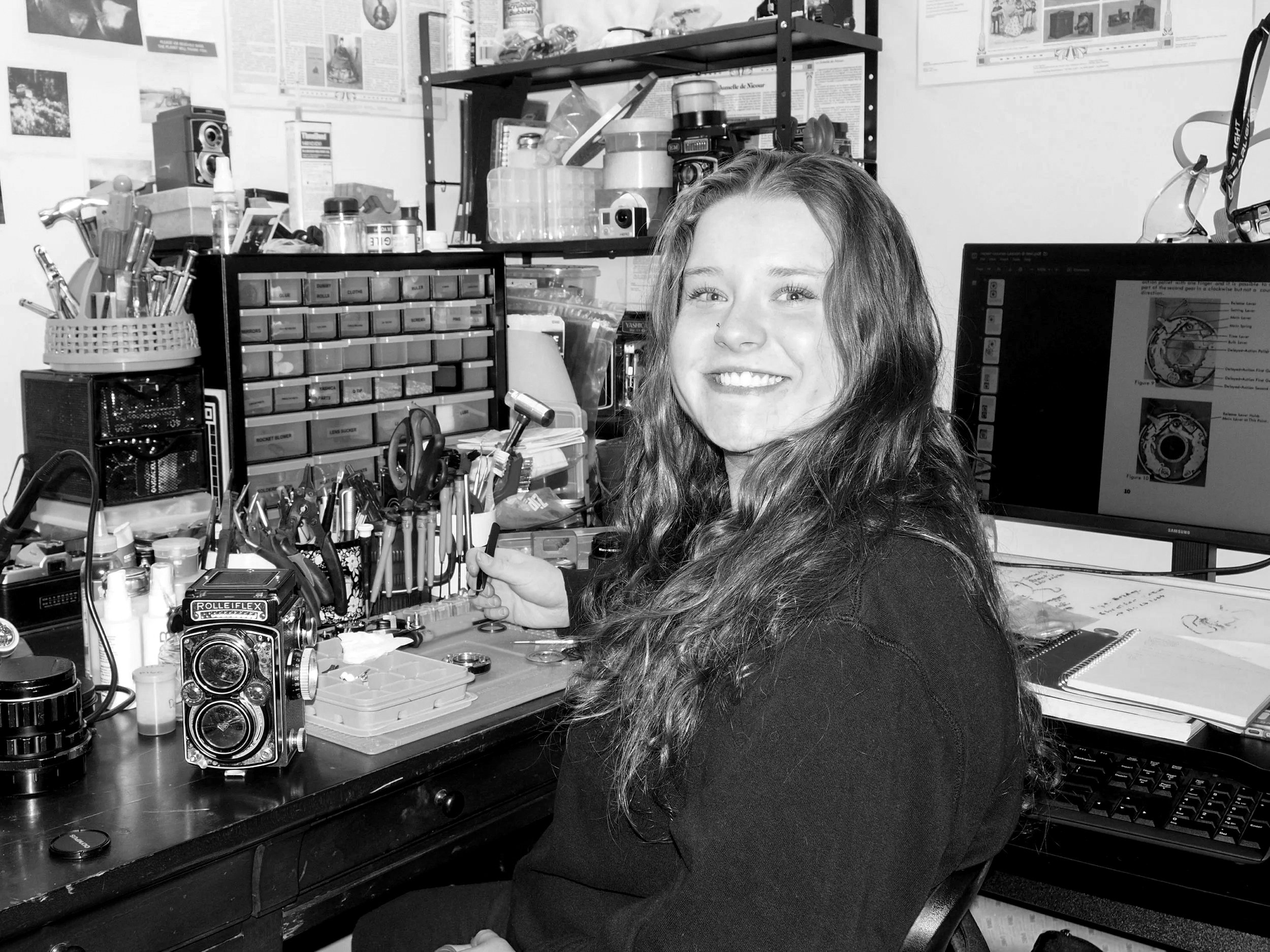 A young woman with long curly hair smiling in a cluttered workshop or electronics lab, surrounded by tools, electronic parts, and a computer monitor.
