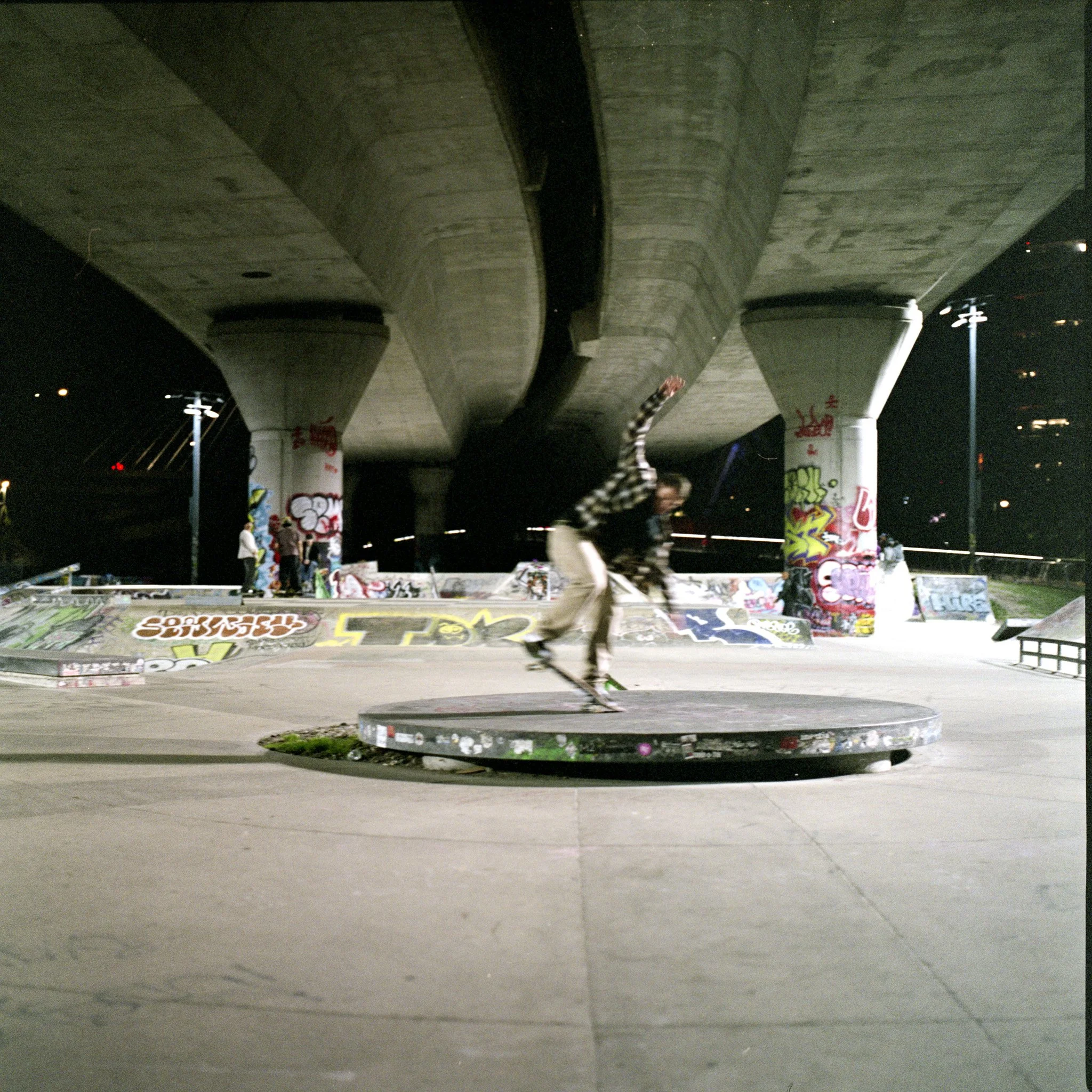 Nighttime view of graffiti-covered pillars supporting an overpass, with a skatepark beneath and a skater performing tricks on a ramp.