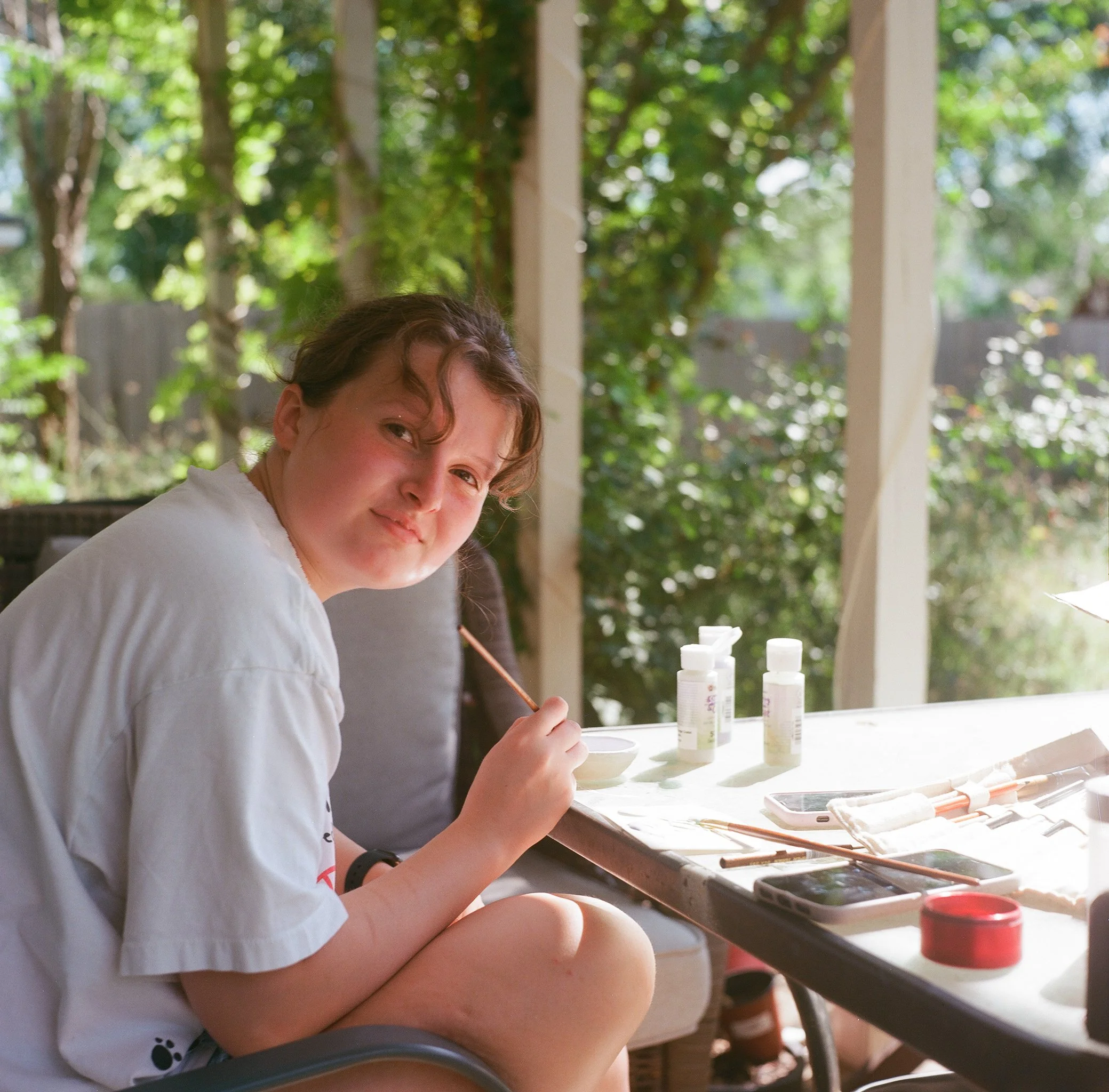 A young girl with brown curly hair and a fair complexion sitting outdoors at a table with art supplies, including bottles of acrylic paints, brushes, and a palette. She is wearing a white T-shirt and has a content expression. The background features lush green trees and a wooden fence, indicating a backyard or garden setting.