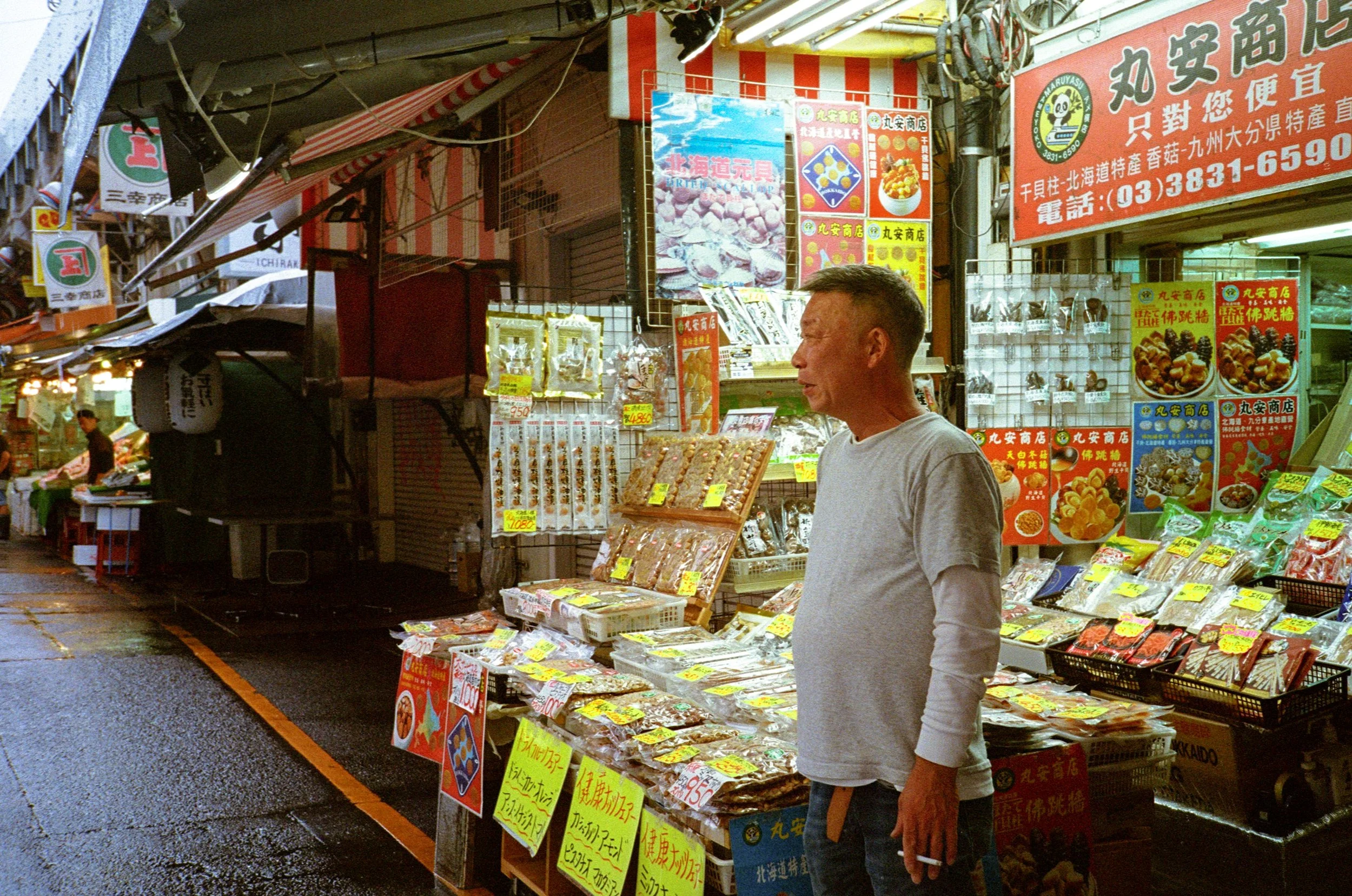 A man stands in front of a colorful outdoor market stall with various packaged goods, posters, and signs in Japanese and Chinese, with a red and white striped canopy overhead.