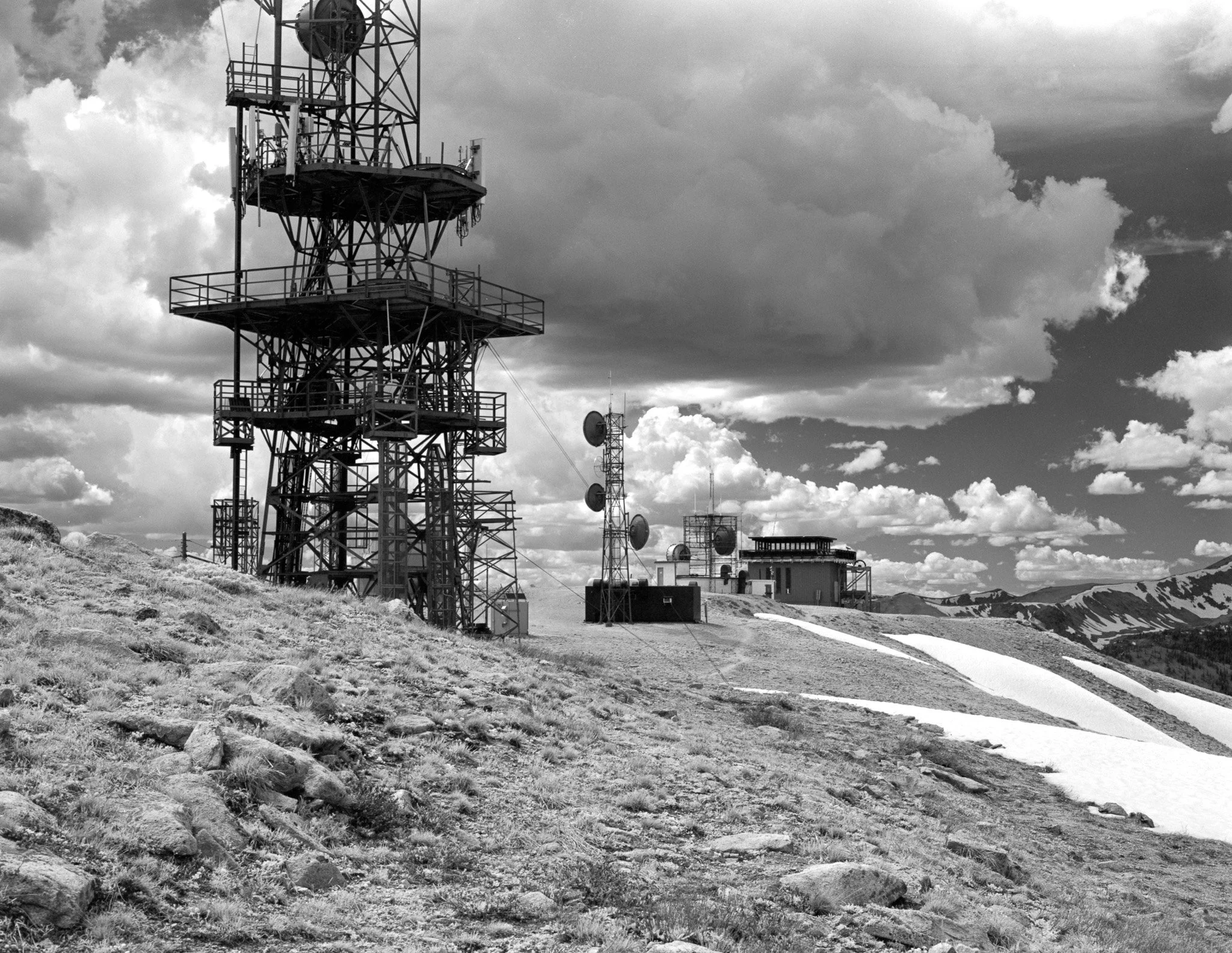 Black and white photo of a tall metal communication tower on a grassy hillside, with a cloudy sky in the background.