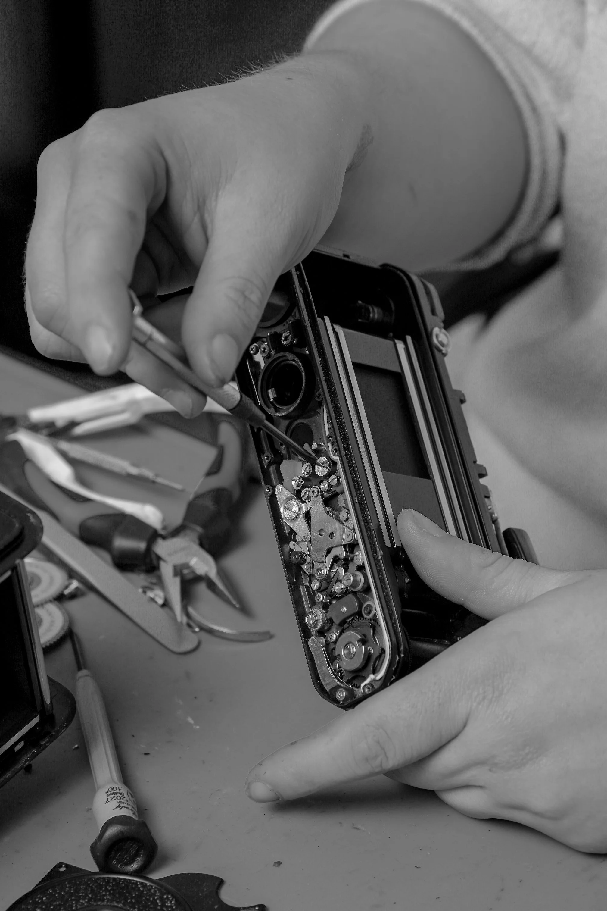 Close-up of a person's hands working on a disassembled camera, with tools and electronic parts scattered on the table.
