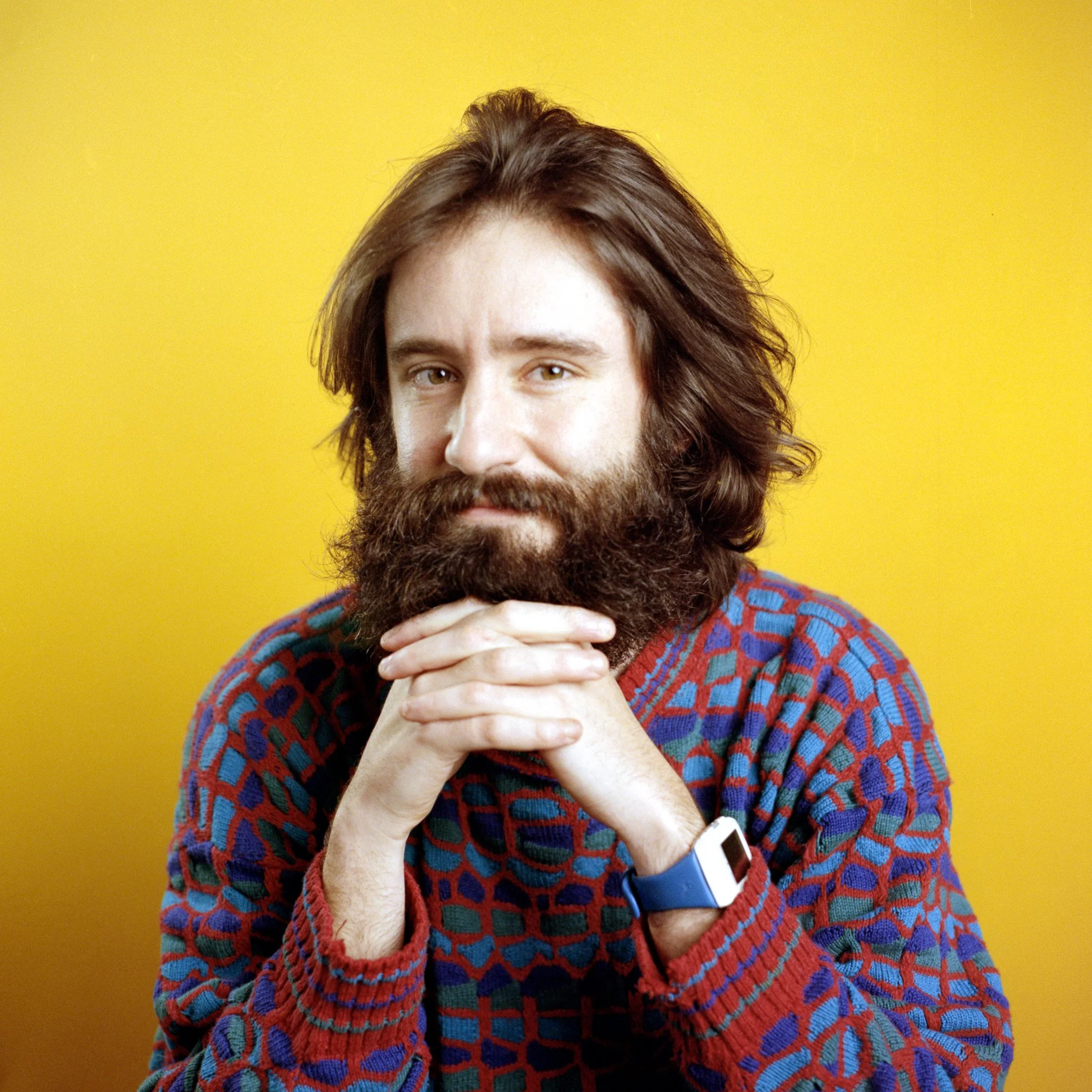 A young man with brown hair and beard, wearing a colorful patterned sweater, sitting against a yellow background, with hands clasped below his chin.