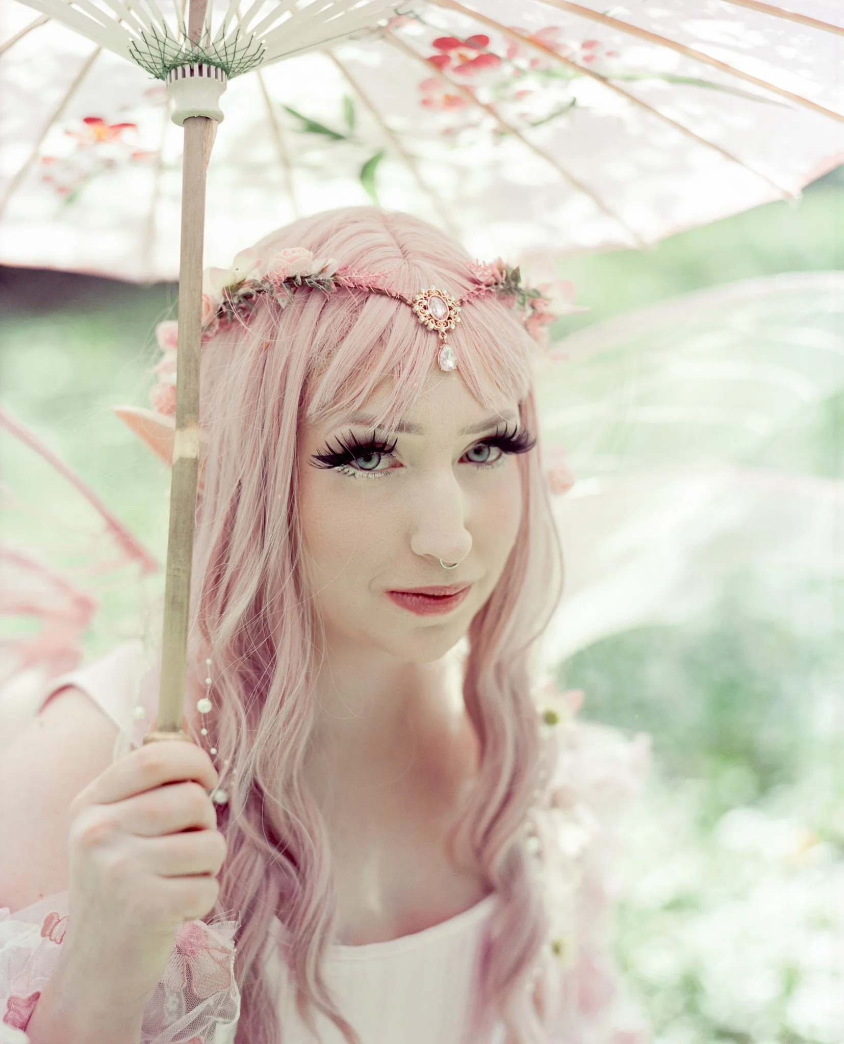A person with long pink hair holding a floral parasol with a pink and white design, outdoors in a garden setting.
