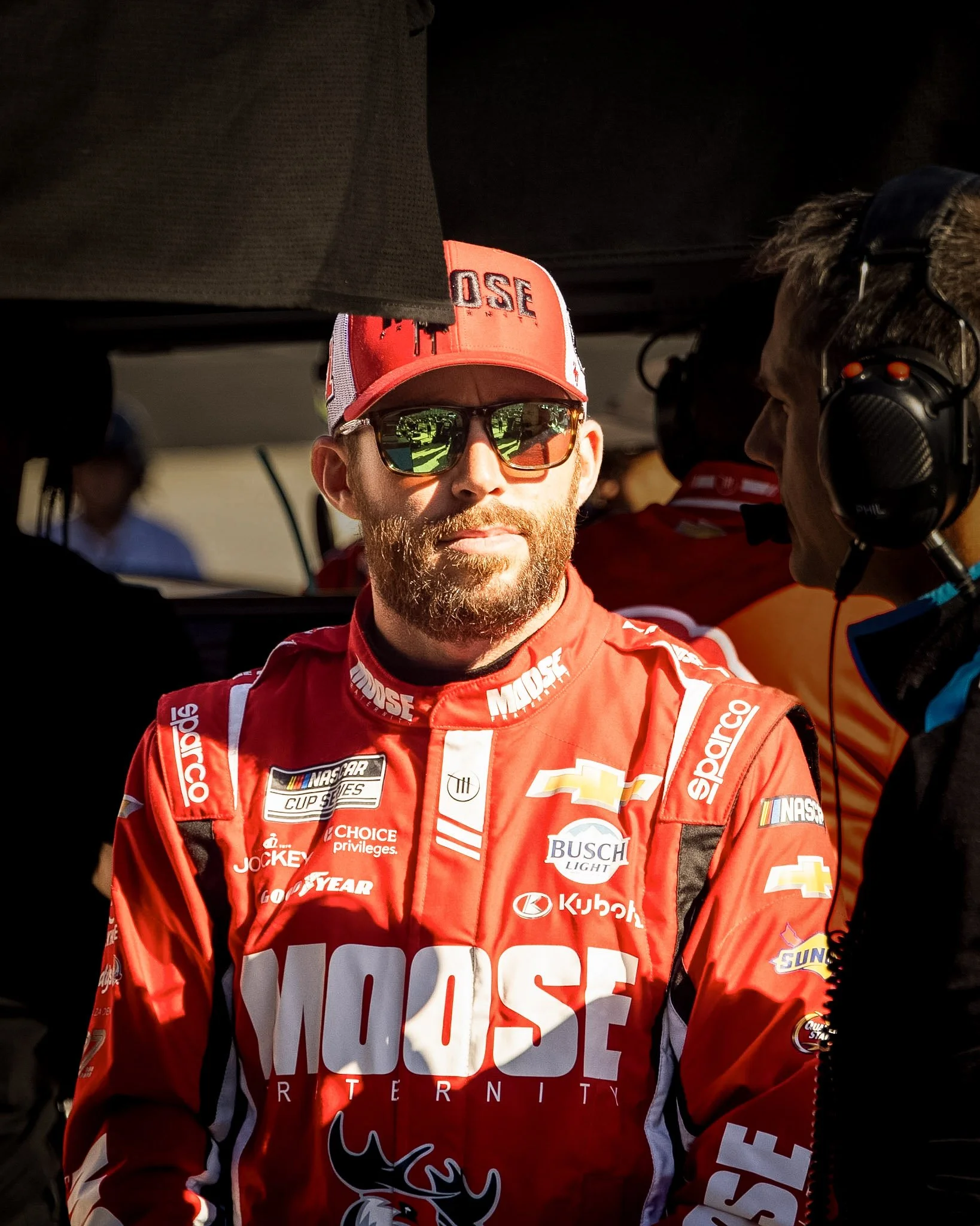 A man wearing sunglasses and a red racing suit with various sponsor logos, standing in a garage or pit area during a racing event.