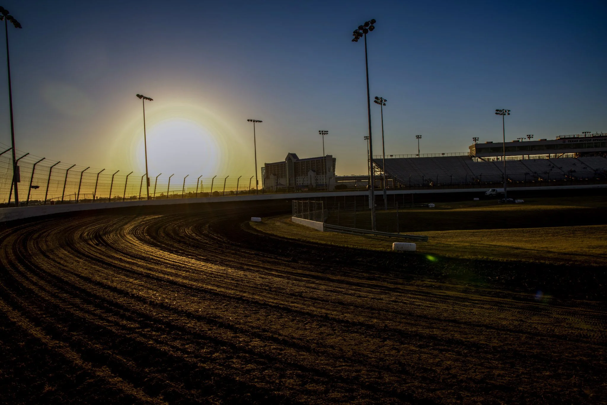 Race track with empty grandstands and tall light poles at sunset or sunrise.