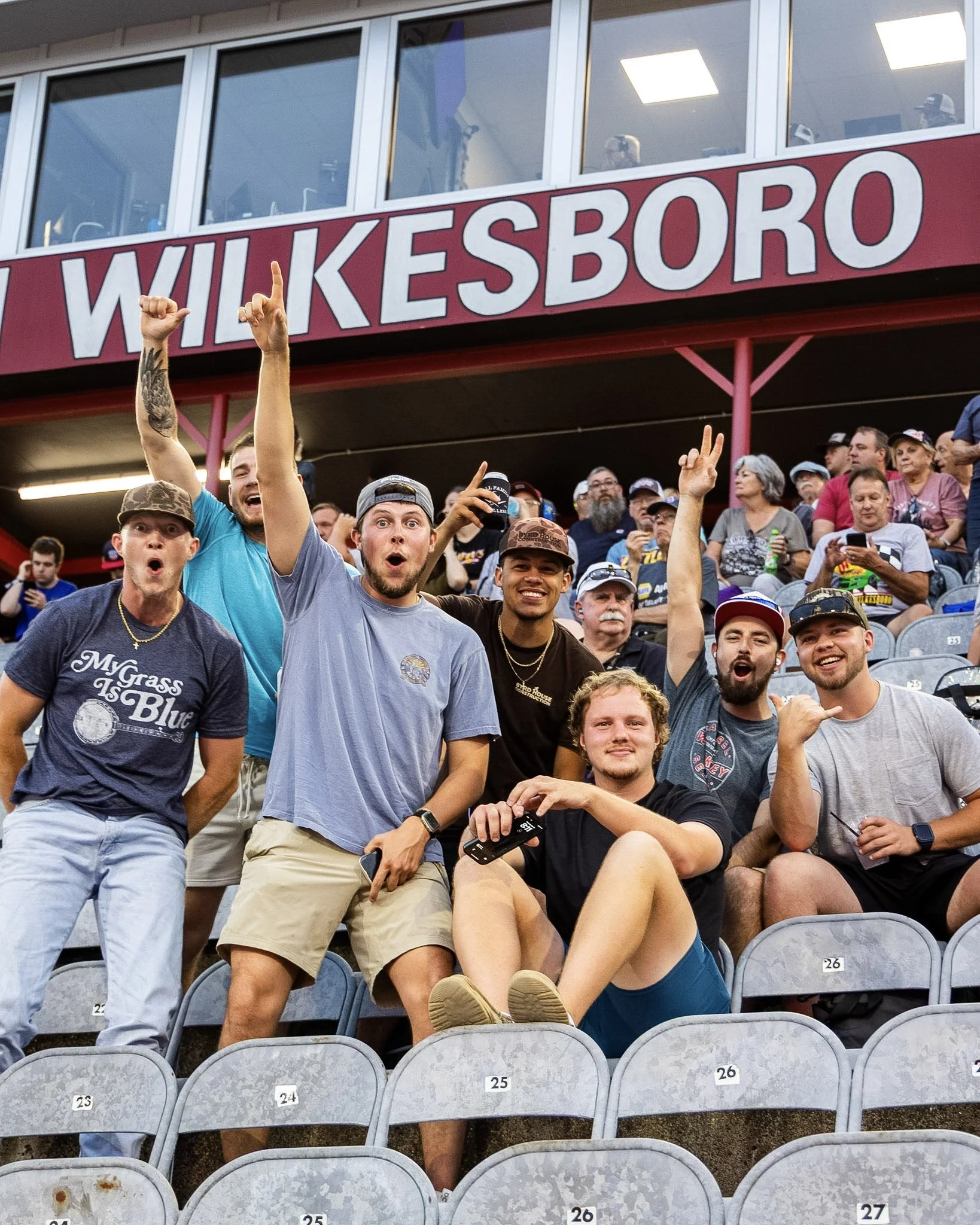 A group of people sitting in stadium seats at a baseball game, with some cheering and holding drinks, while others are looking at the camera or their phones. The stadium has a sign that reads 'WILKESBORO' and numbered seats.