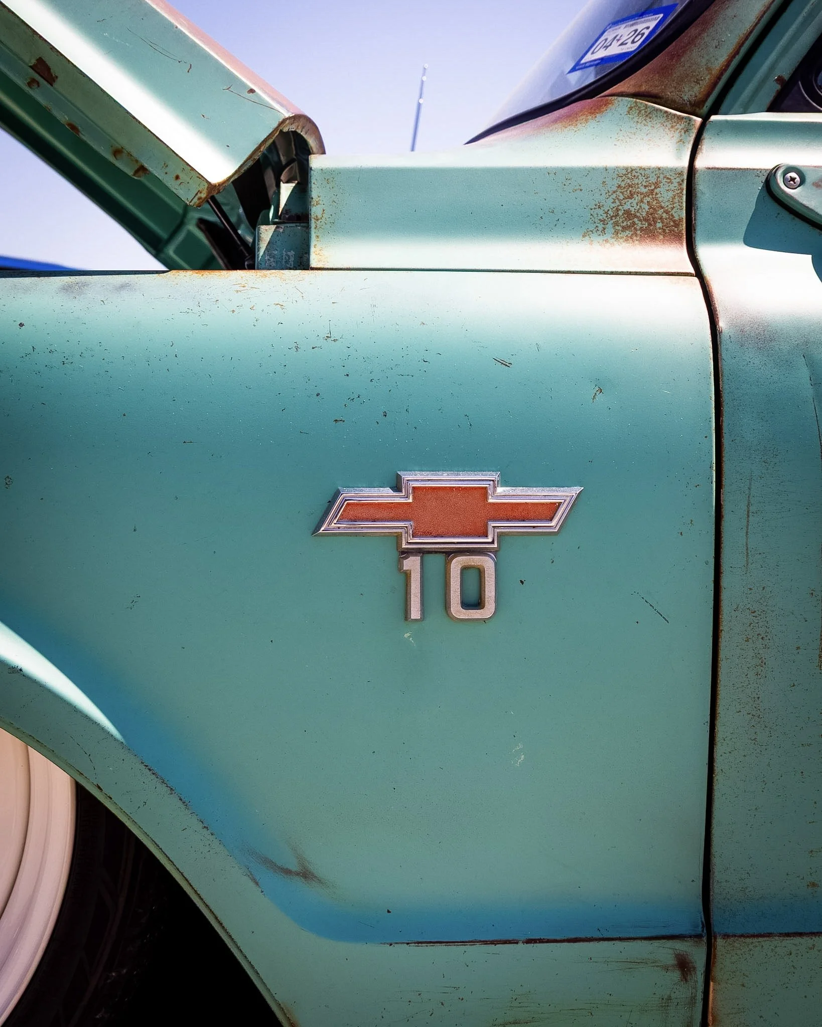 Close-up of an old turquoise pickup truck with rust spots and a badge that reads '10', part of a Chevrolet logo, on the side.