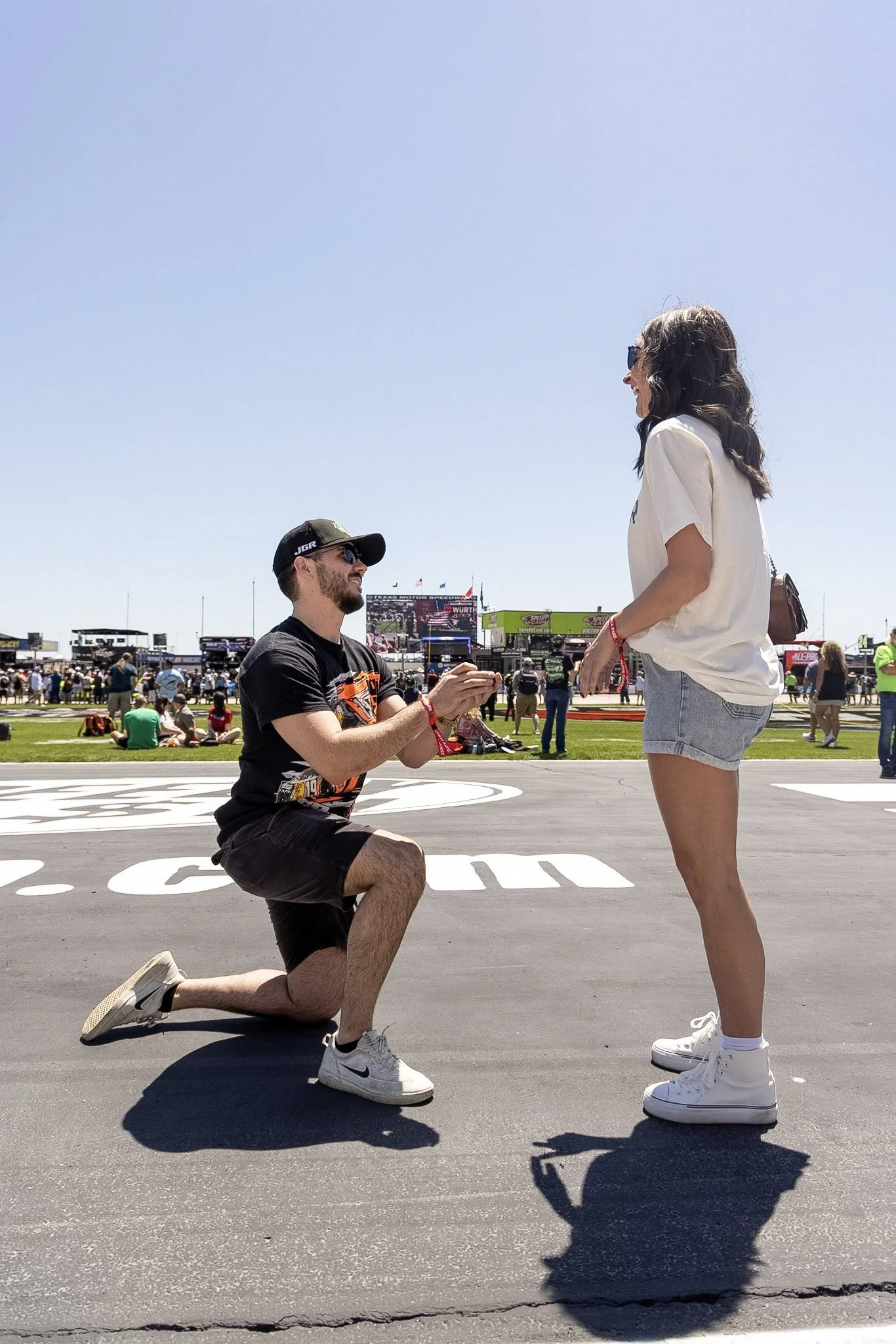 A man is kneeling on one knee, proposing to a woman who is standing in front of him at a race track. The man is wearing a black t-shirt, shorts, white sneakers, sunglasses, and a cap. The woman is smiling, wearing sunglasses, a white t-shirt, denim s