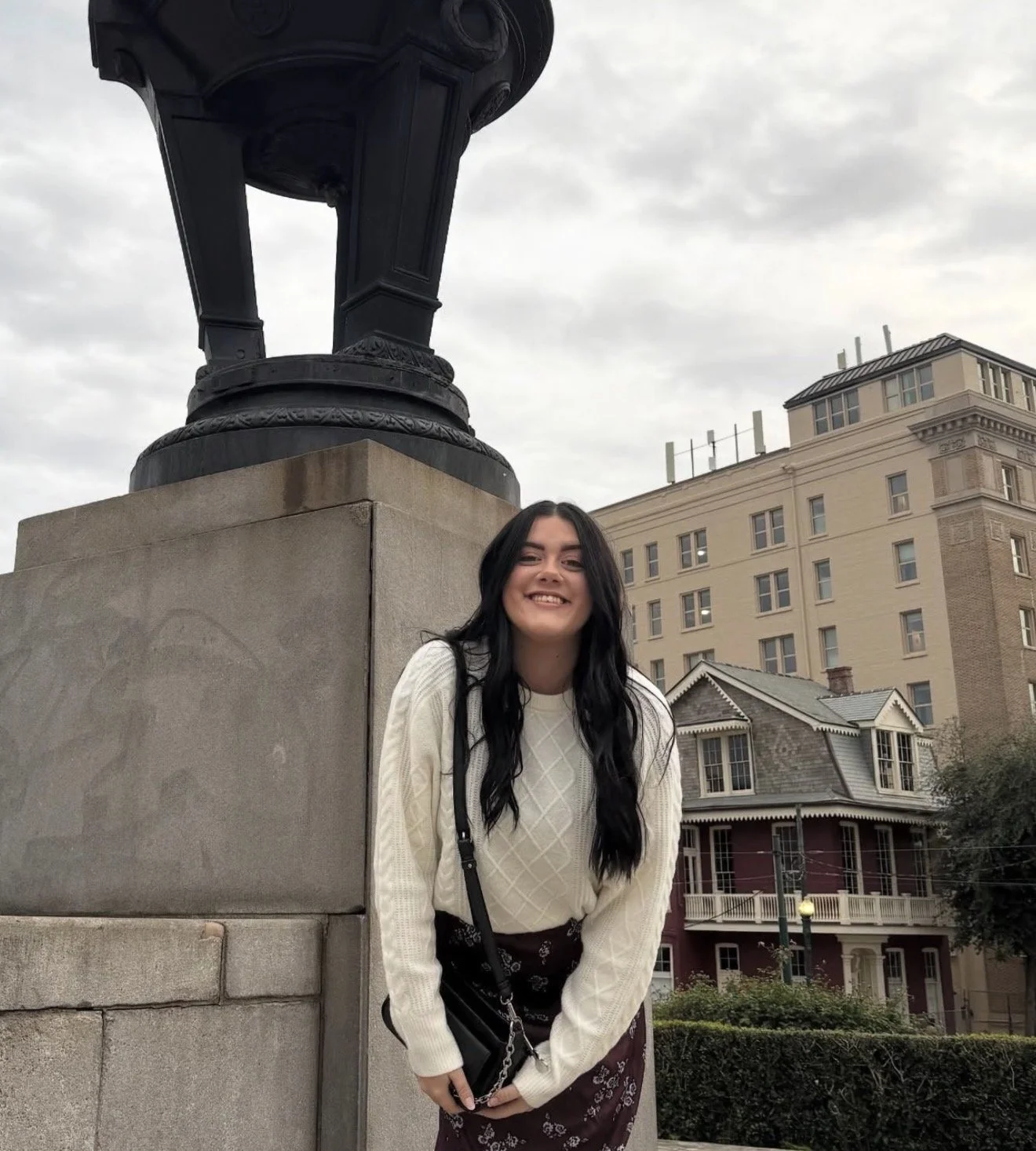 A young woman with long black hair smiling, standing outdoors near a stone monument base with a large, ornate black statue on top, and a cityscape with multi-story buildings and a house in the background.