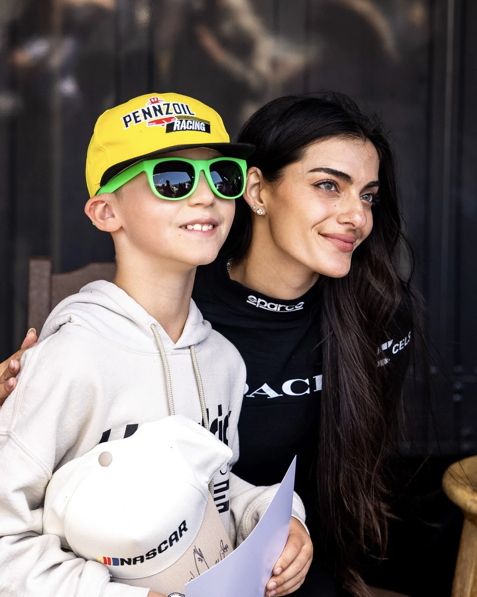 A young boy wearing a yellow cap and bright green sunglasses is standing next to a woman with long dark hair, both smiling. The boy is holding a piece of paper and has on a white hoodie with racing-related logos. The woman is dressed in a black shirt