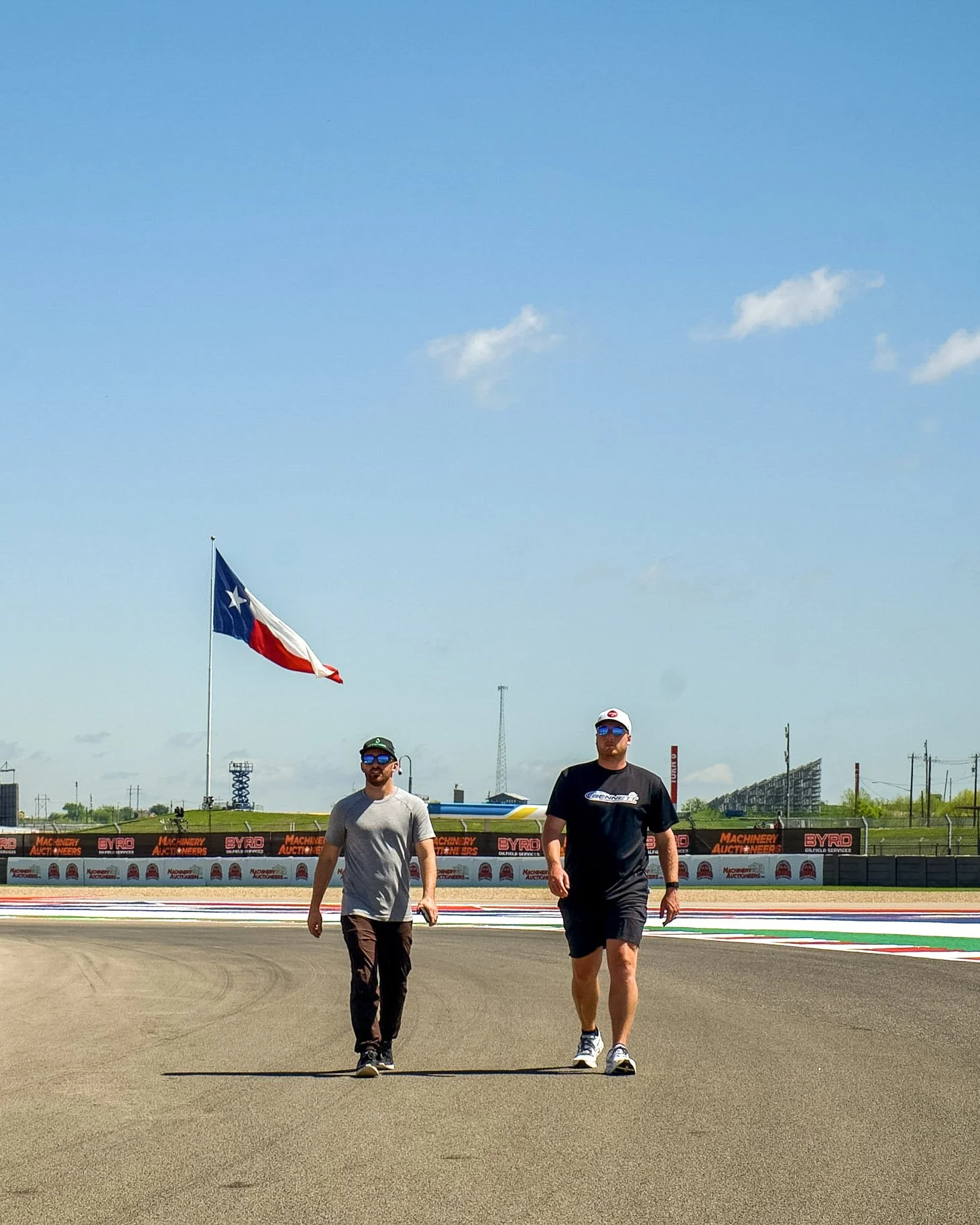 Two men walking on a race track with an American flag in the background, a clear blue sky overhead, and some spectators and structures in the distance.