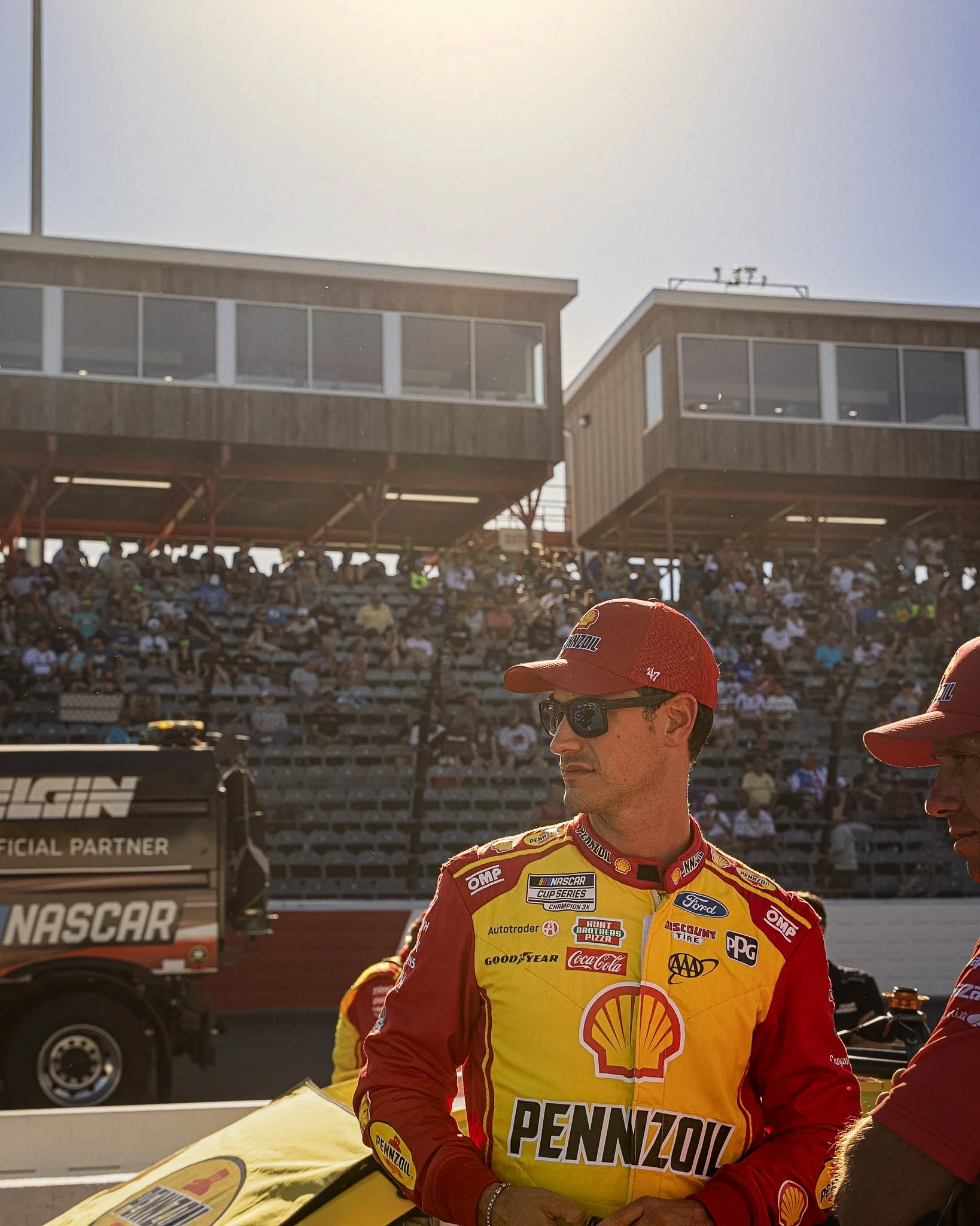 Race car driver in yellow and red racing suit with sponsorship logos, standing near his yellow race car overhang, with a crowd and grandstands in the background under a clear sky.