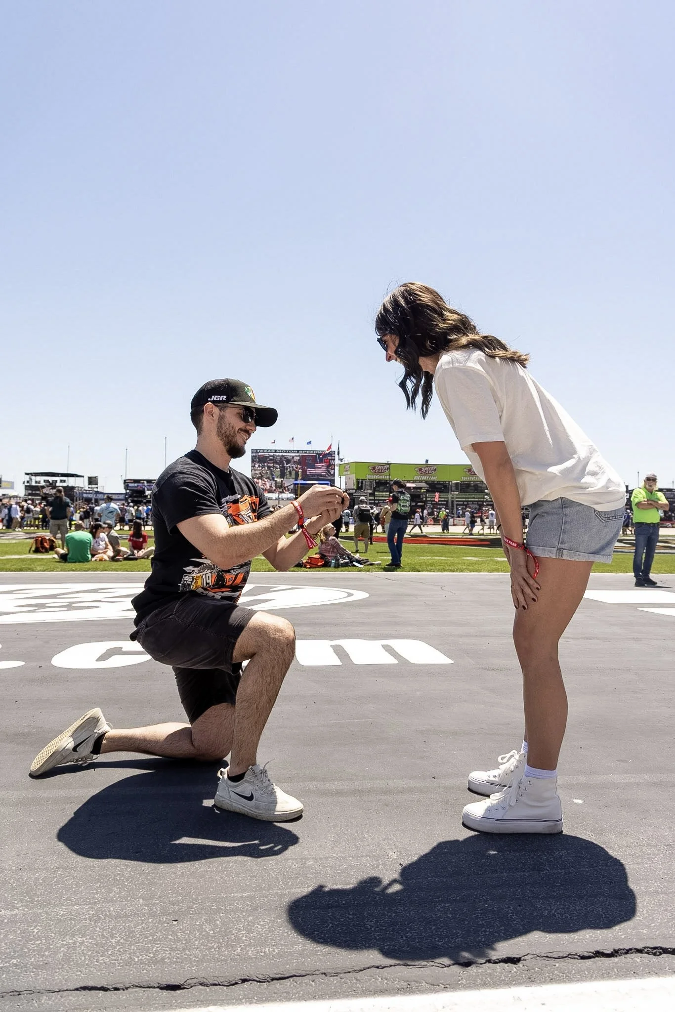 A man kneeling on one knee in front of a woman, holding her hand, while she bends forward slightly at a racing event or outdoor festival. The man wears a black cap, sunglasses, black shirt, and shorts. The woman wears a white t-shirt, gray shorts, an