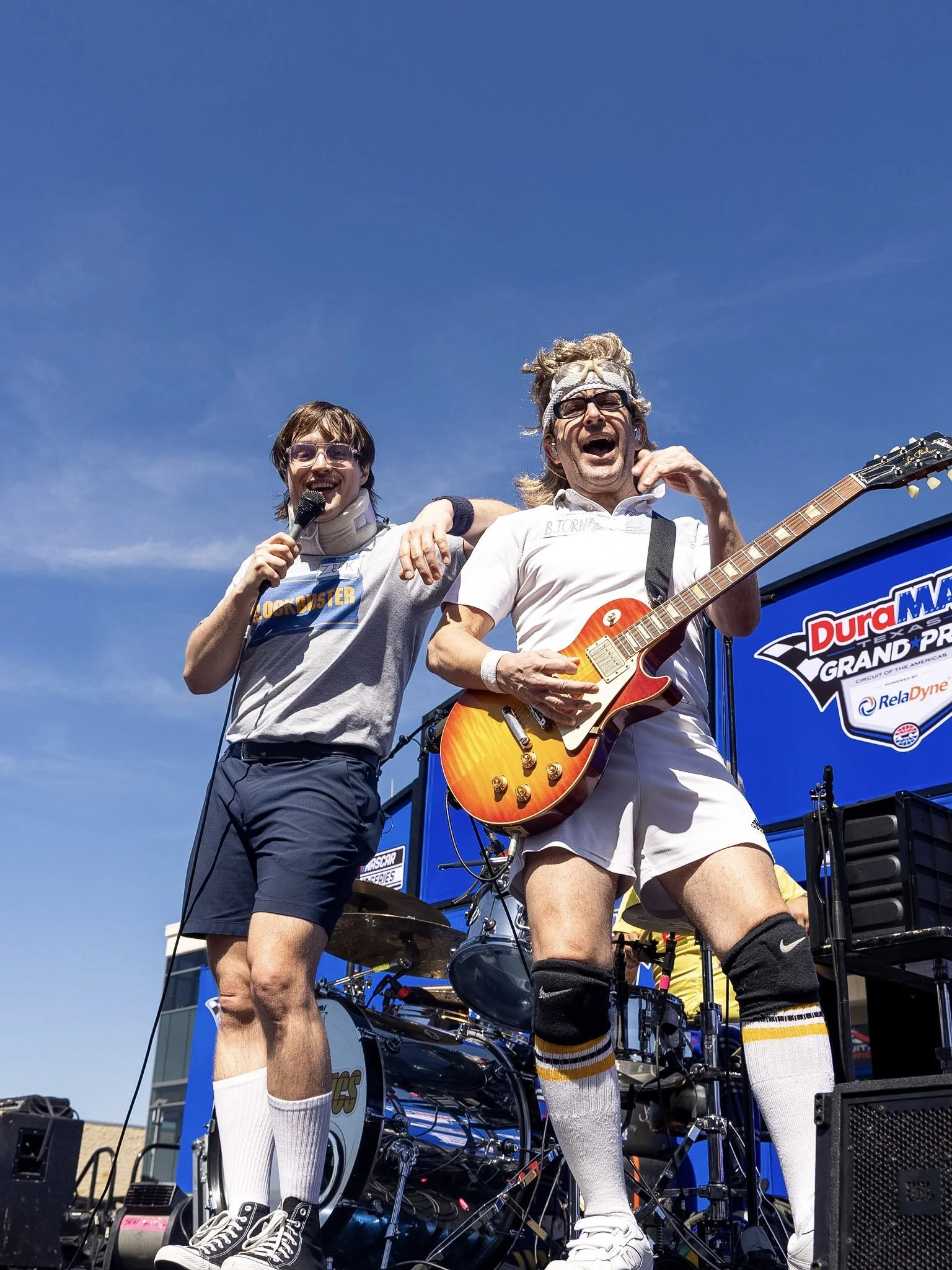 Two men wearing athletic gear, one playing guitar and the other holding a microphone, performing on stage outdoors against a blue sky at a racing event.
