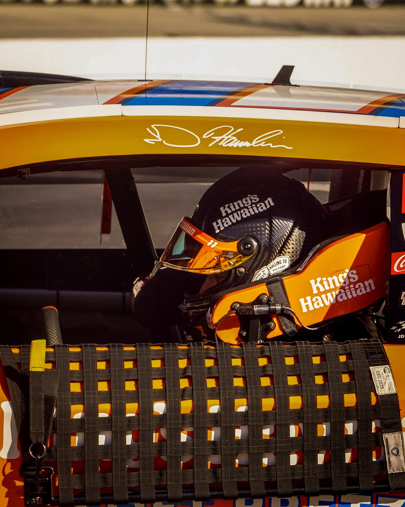 Close-up of a race car driver wearing a helmet and a racing suit, with a race car's rear wing and a signed autograph visible in the background.