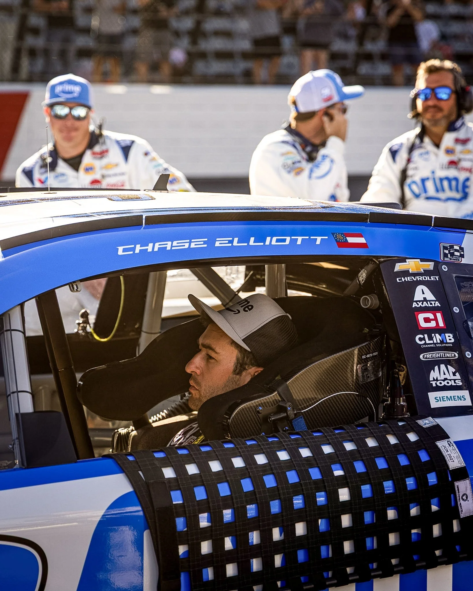Close-up of a race car driver inside his race car, with three crew members standing behind the car wearing white uniforms and caps, with a blurred background of trees and a wall.