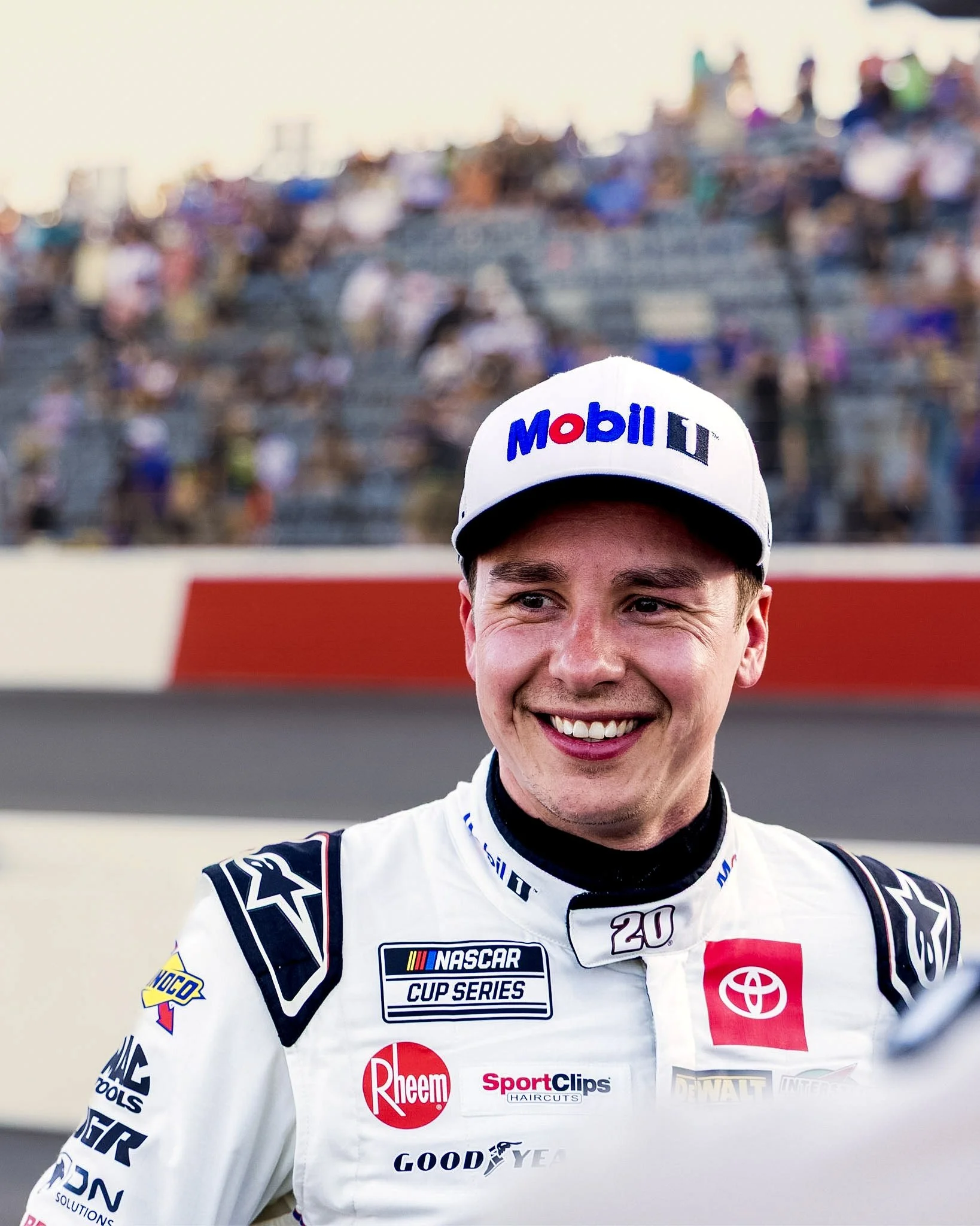 Close-up of a smiling race car driver wearing a white racing suit with sponsor logos, including NASCAR Cup Series, Toyota, and Rheem, and a white cap with the Mobil 1 logo, standing in front of a blurred grandstand filled with spectators.