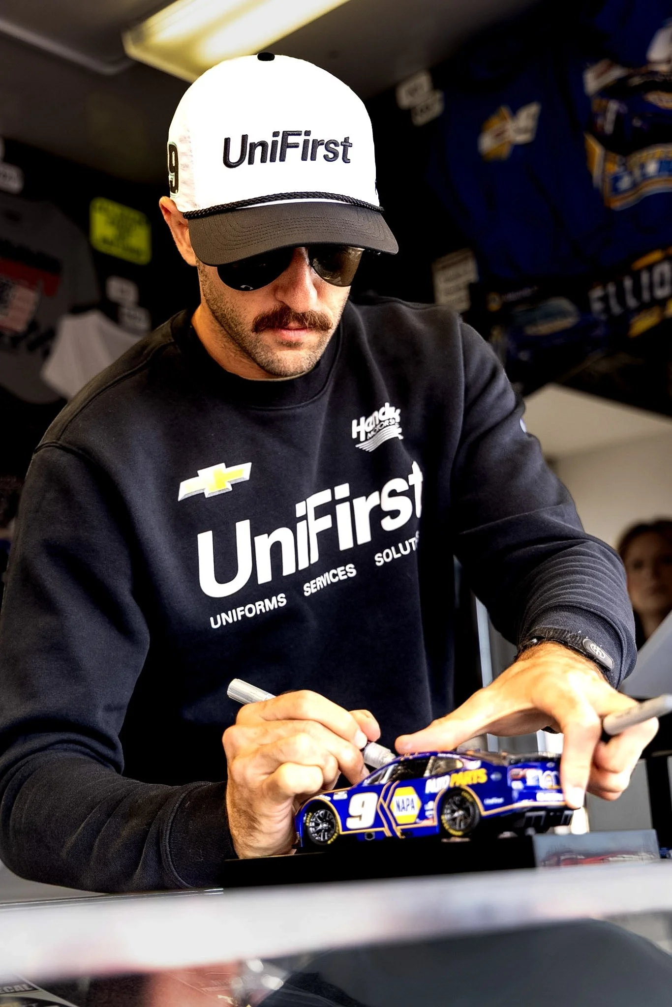 A man wearing sunglasses and a cap with 'UniFirst' text signs a miniature racing car at a table. He is wearing a black 'UniFirst' branded shirt.