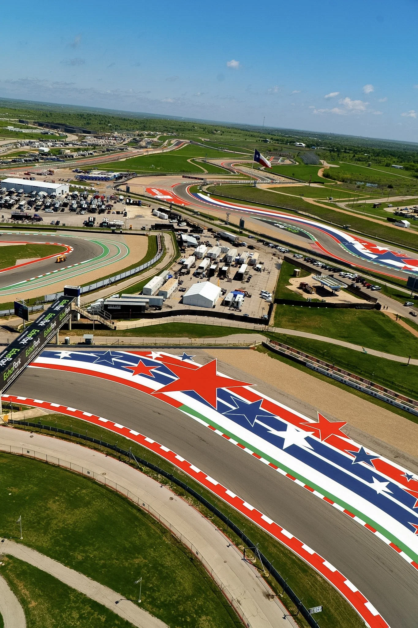 Aerial view of a race track with colorful painted markings including stars, red, white, and blue stripes, and various lanes. The track is surrounded by parking lots, tents, and a large flagpole with the Texas state flag. Green grass and trees enclose