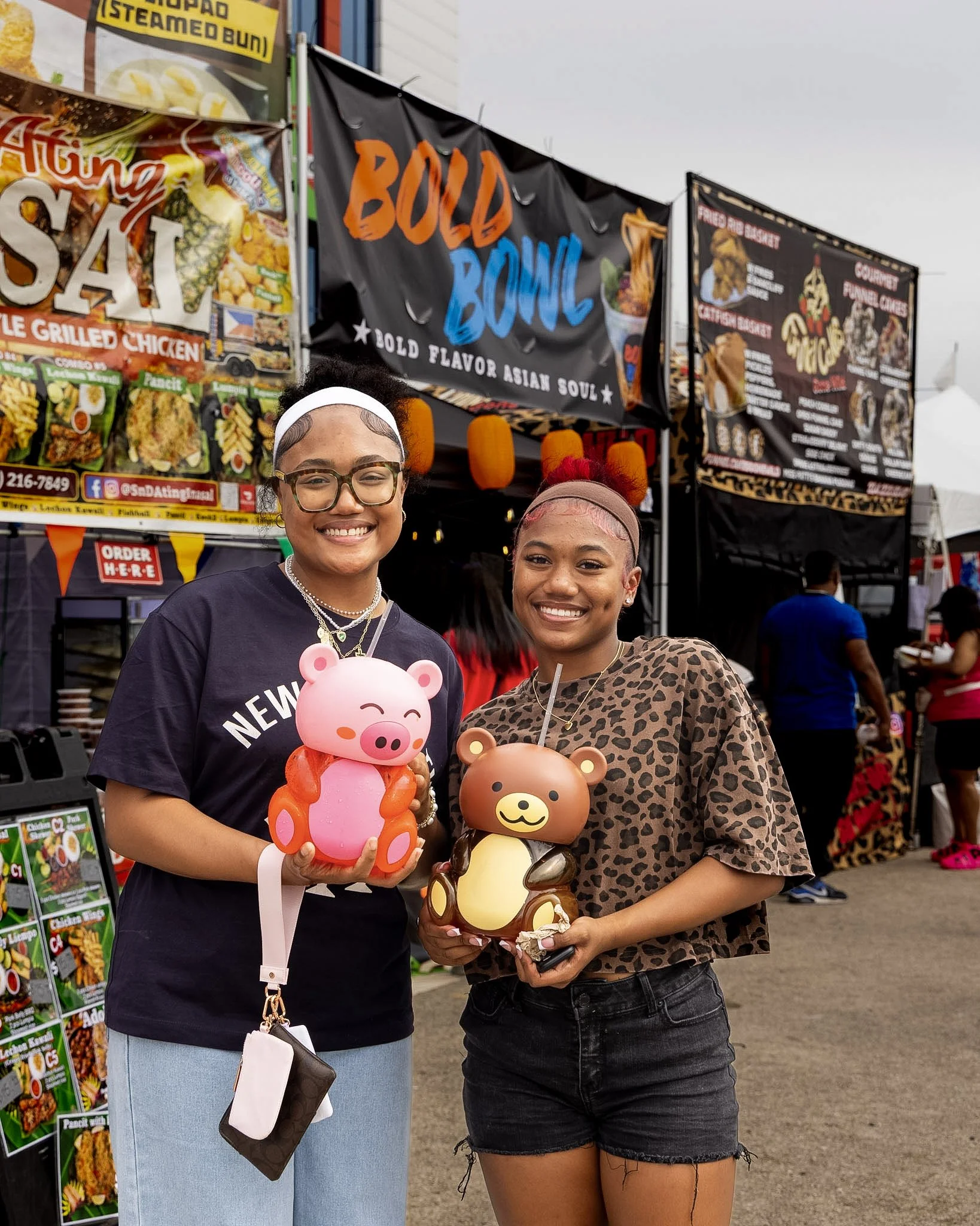 Two young women standing in front of food vendor stalls at an outdoor event, holding pig-shaped and bear-shaped desserts, smiling at the camera.