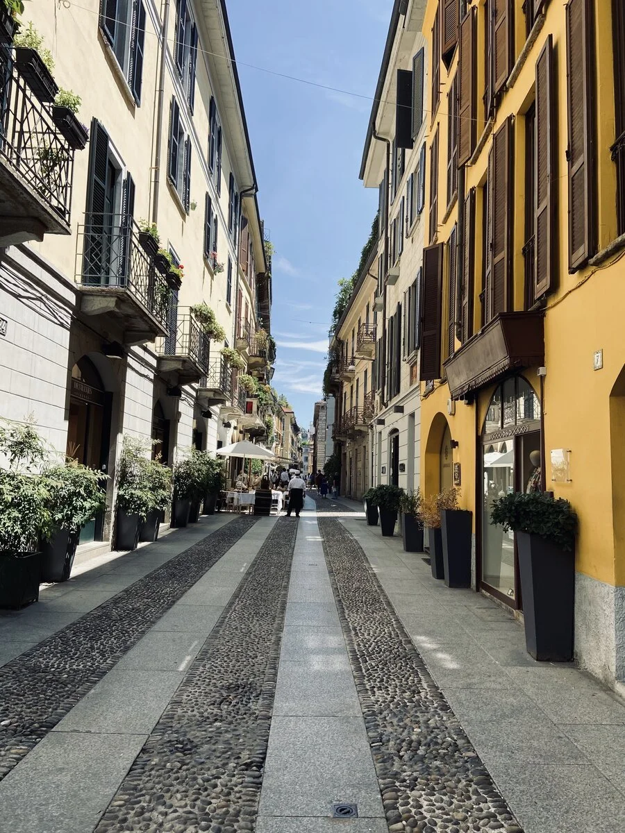 Strada pedonale tra edifici con negozi e ristorante, vasi di piante ai lati, sotto un cielo azzurro con poche nuvole.