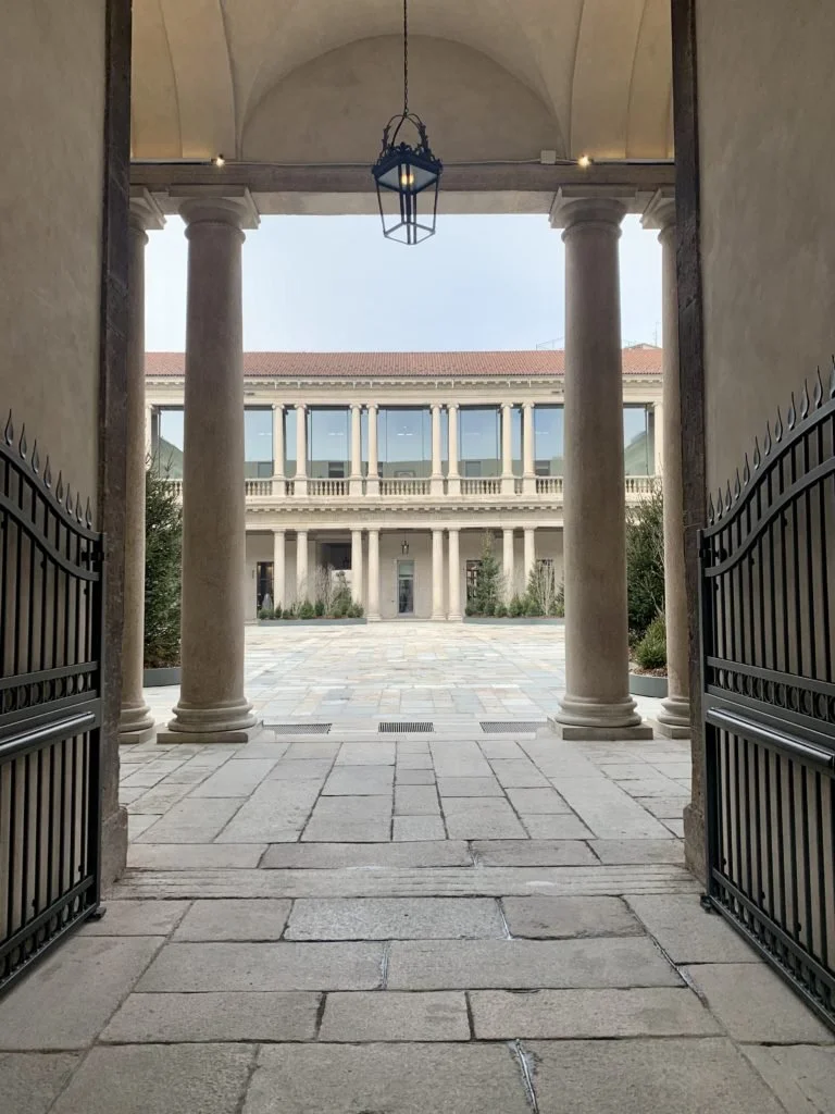 Vista attraverso un arco in stile classico con colonne di pietra su un cortile con pavimentazione in pietra, che mostra un edificio con colonne e balconi in stile classico.