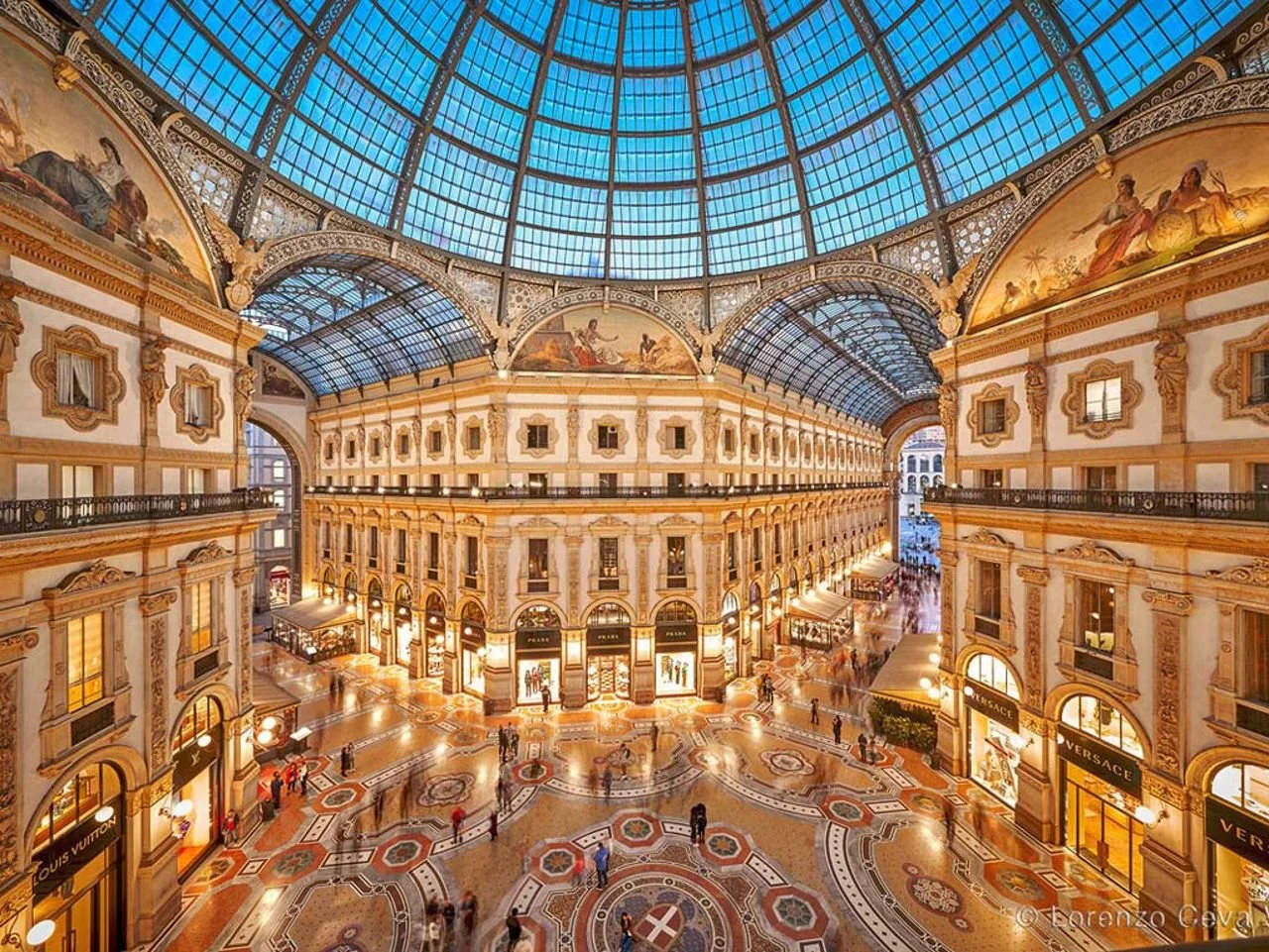 Interno di un grande centro commerciale di lusso con decorazioni dorate, negozi di stilisti come Louis Vuitton, Versace, e un grande soffitto di vetro che lascia entrare la luce naturale.