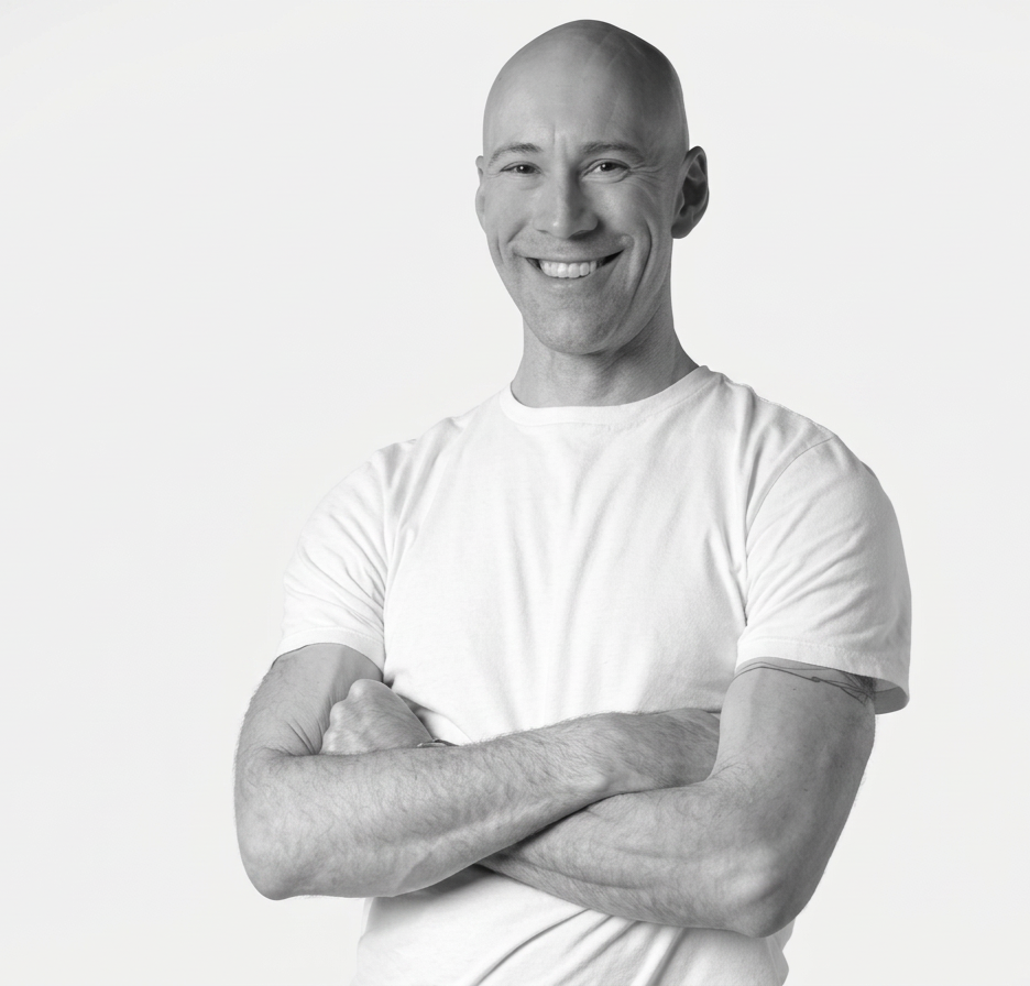 Black and white photo of a smiling man with a shaved head, wearing a white t-shirt with arms crossed against a plain background.