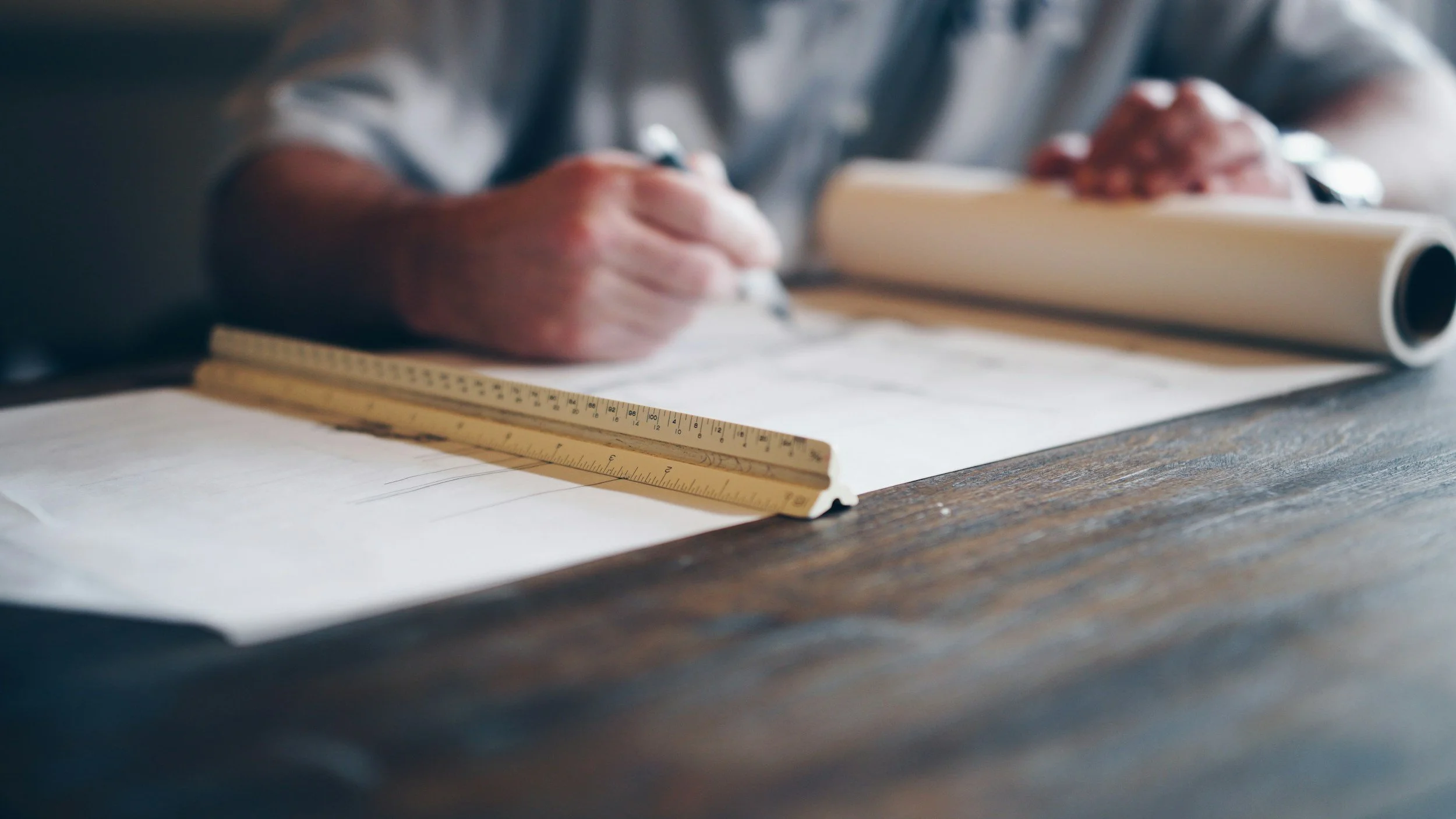 Close-up of a person's hand writing on paper with a pencil, a ruler, and a rolled-up blueprint on a wooden table.