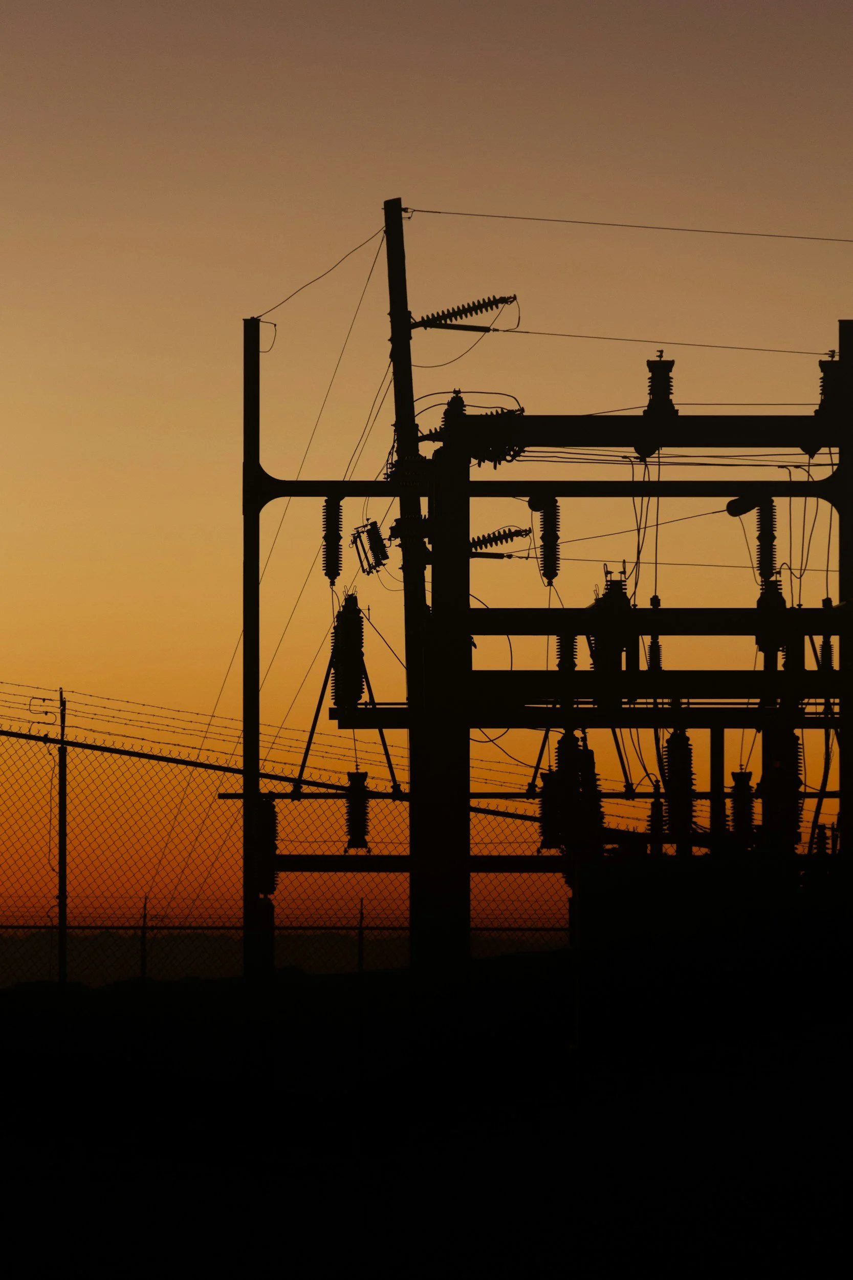 Silhouettes of electrical power lines and transformers against a sunset or sunrise sky with a chain-link fence in the foreground.