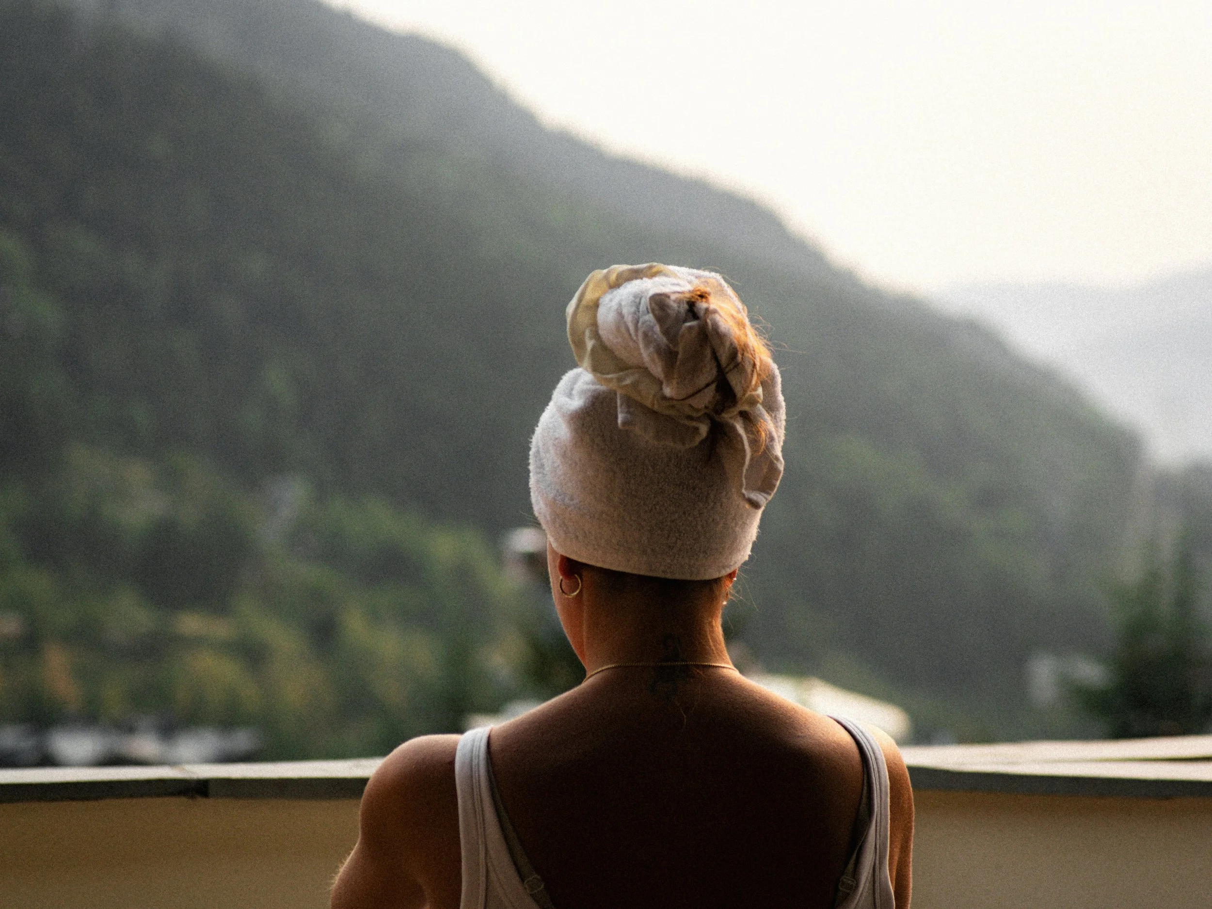 A woman with short hair tied in a bun, wearing a white tank top and hoop earrings, standing outdoors and looking at mountains in the distance during sunset or sunrise.