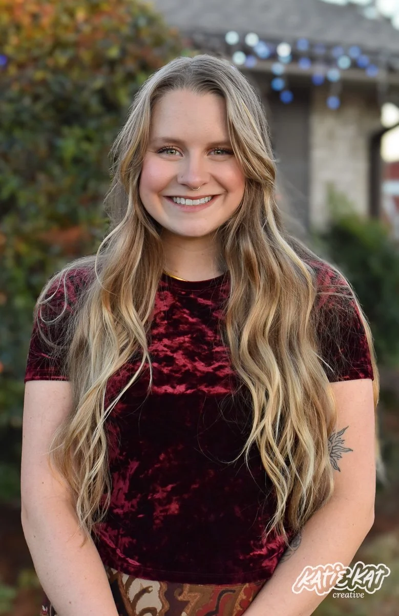 A young woman with long wavy blonde hair, smiling, standing outdoors in front of green shrubbery and a house with string lights.
