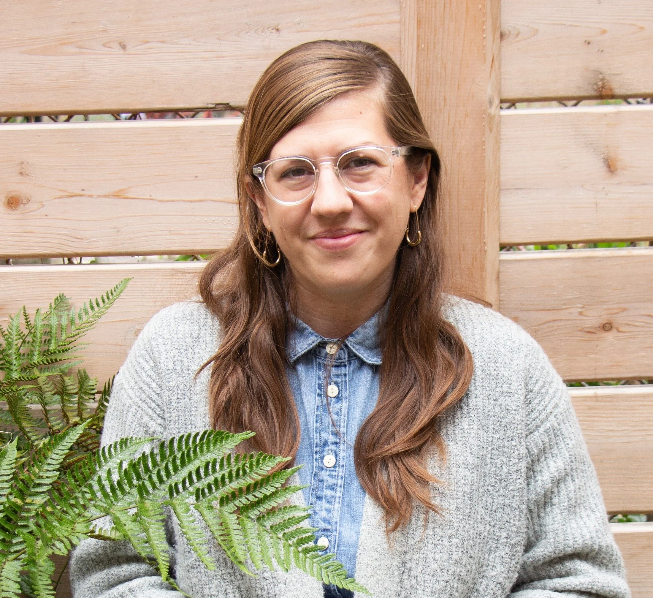 A female therapist with long brown hair, wearing glasses, a denim shirt, and a gray sweater, standing in front of a wooden fence with green plants in the foreground.