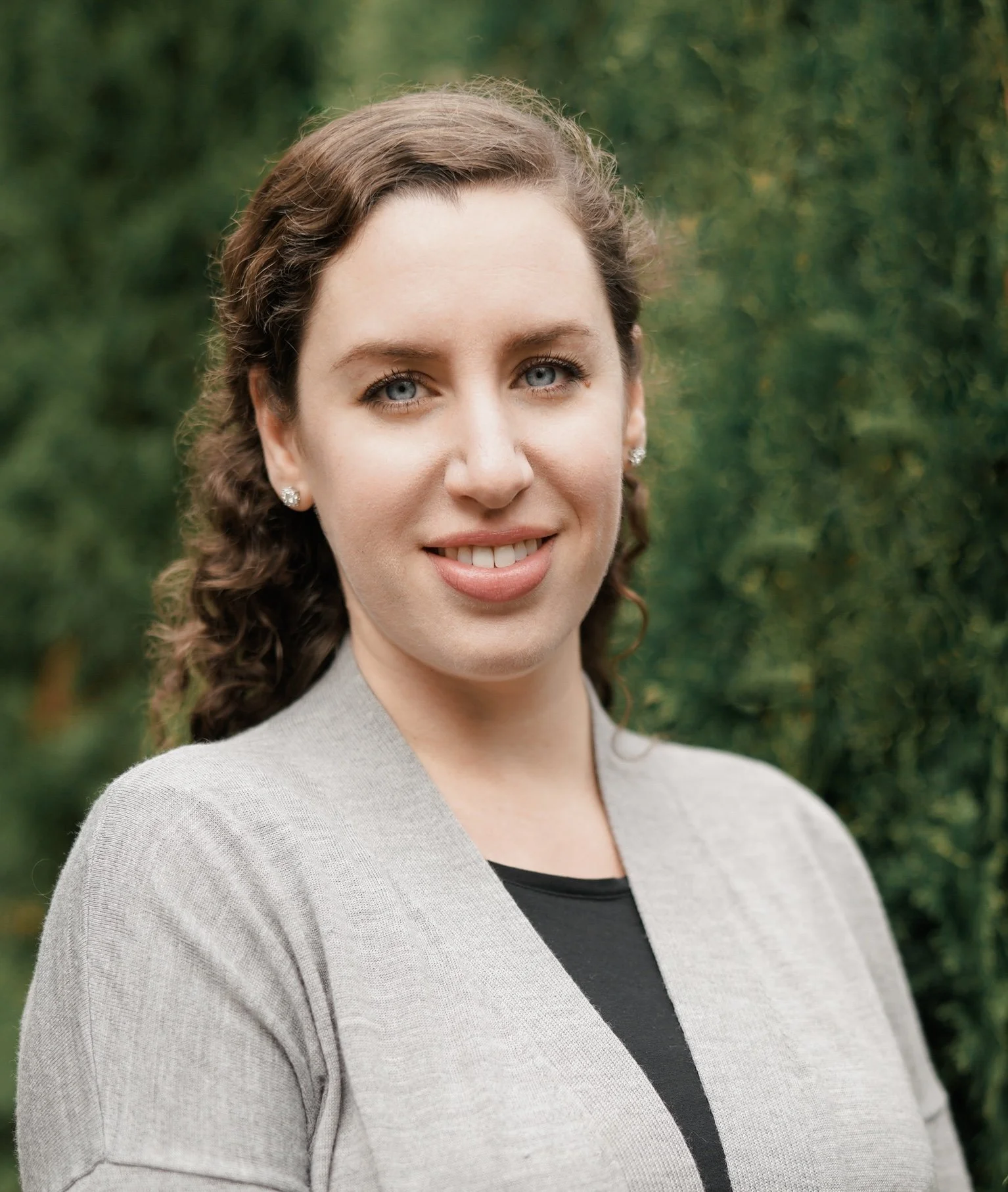 Close-up of a female therapist with curly brown hair, blue eyes, and earrings, wearing a light gray blazer and black top, outdoors with greenery in the background.