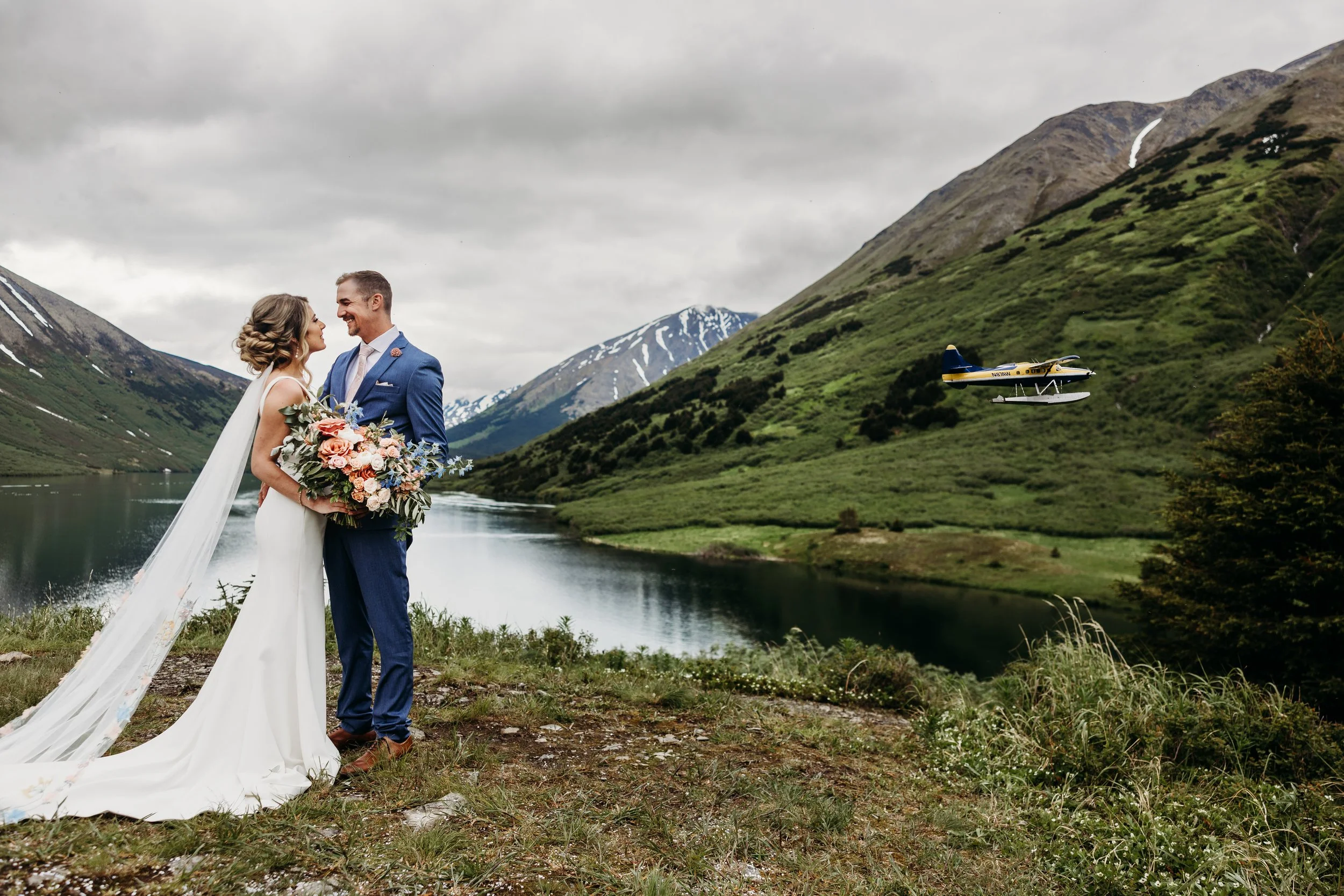 A bride and groom standing by a lake in a mountainous landscape, with a seaplane flying nearby.