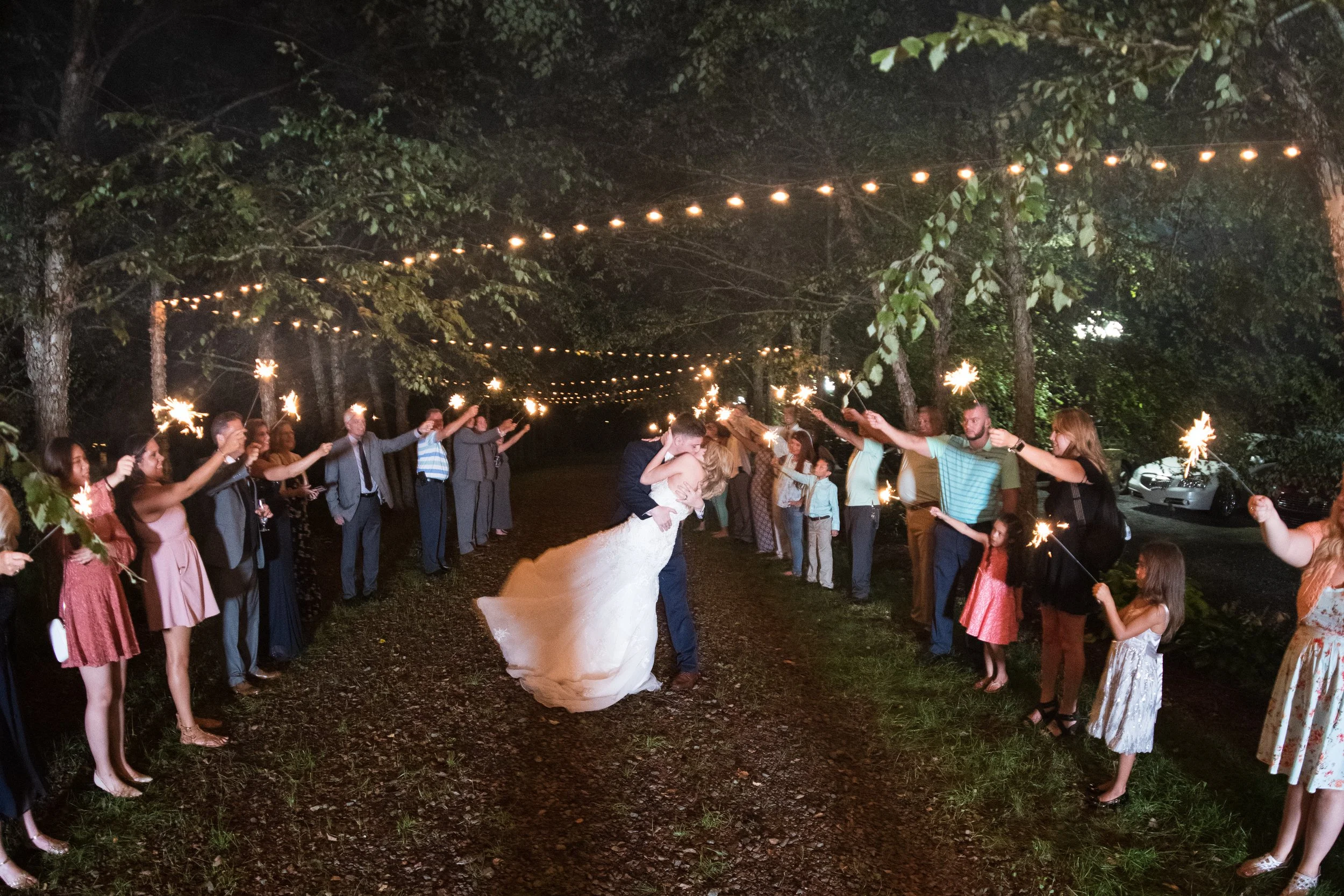 People gather outdoors at night holding sparklers and celebrating, with string lights overhead and trees in the background. The bride and groom are dancing in the center.