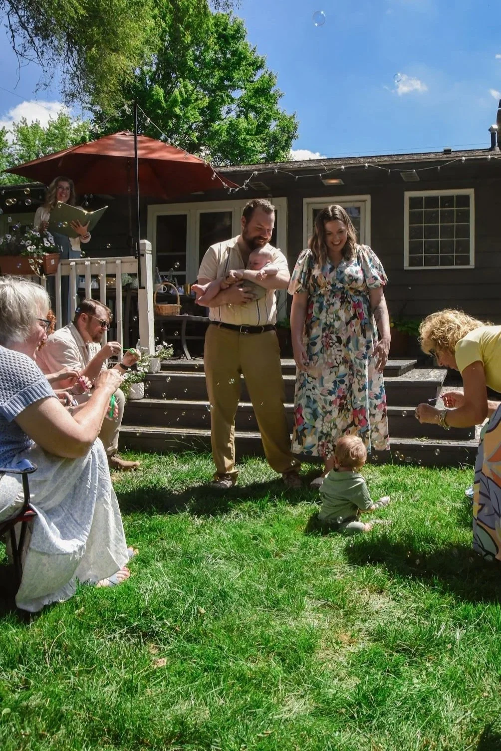A backyard gathering with adults and a baby sitting on the grass, blowing bubbles and enjoying a sunny day.