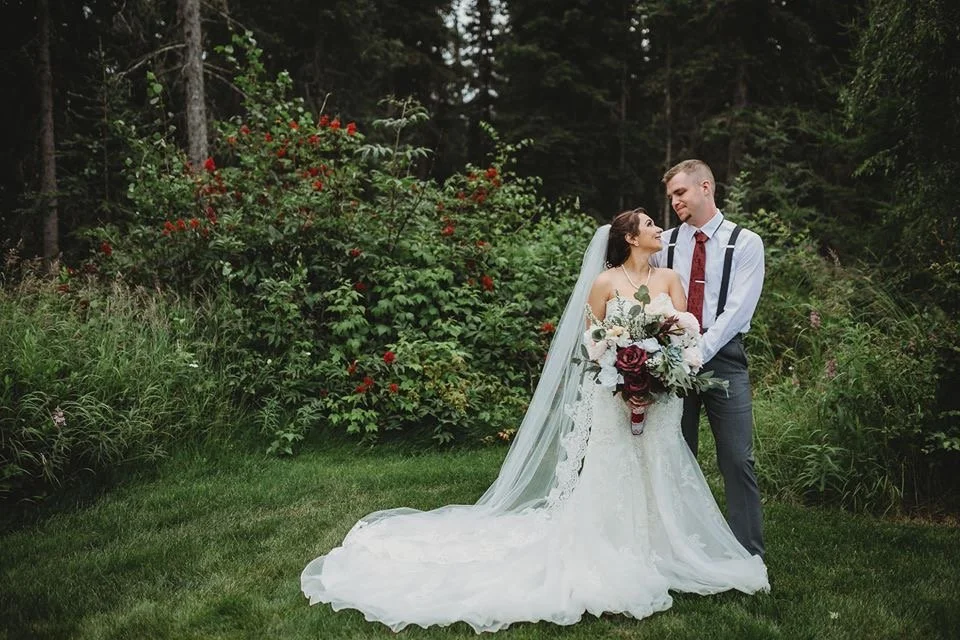 Bride and groom stand close together outdoors in a garden, gazing at each other. The bride wears a white wedding gown with a long train and veil, holding a large bouquet of flowers. The groom wears a white shirt with suspenders, a red tie, and gray trousers. Behind them are lush green trees and red flowering bushes.