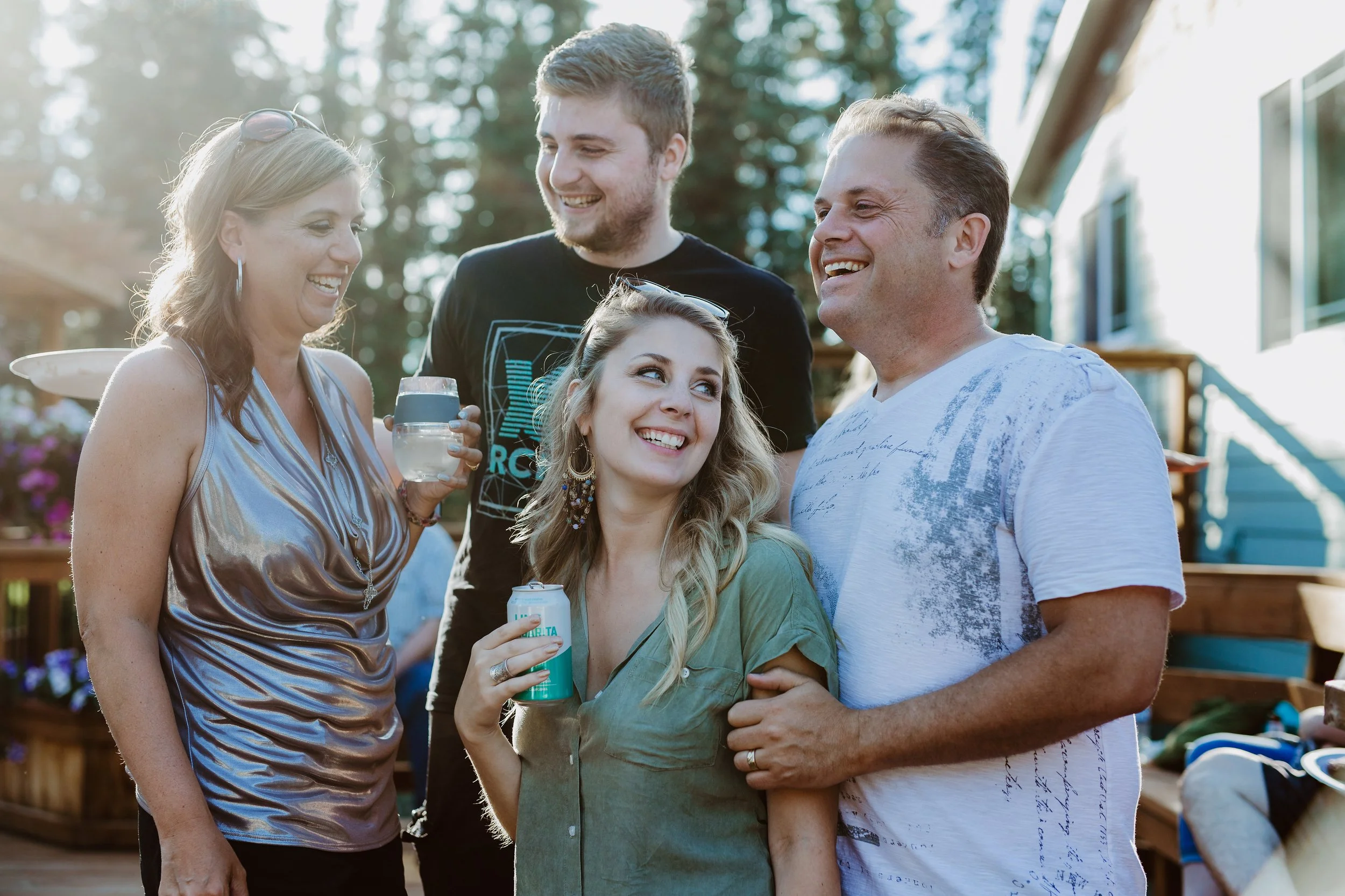 Group of five friends smiling and laughing outdoors during daytime, some holding drinks, in a backyard setting with trees and a house.