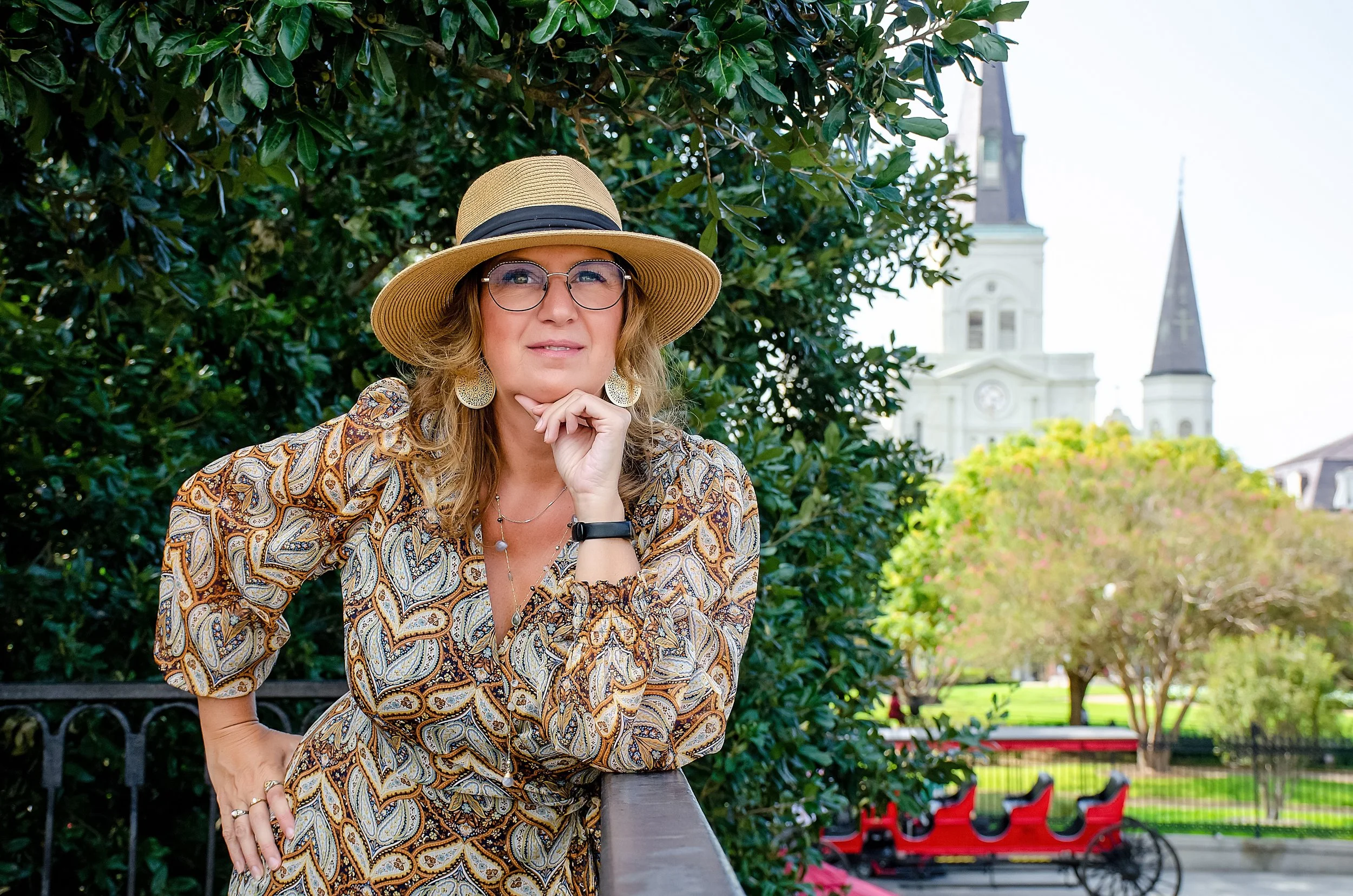 Carrie, Humanist Celebrant and founder of All Things Joyful, standing outdoors in a patterned dress and straw hat, with a historic cathedral and lush greenery in the background