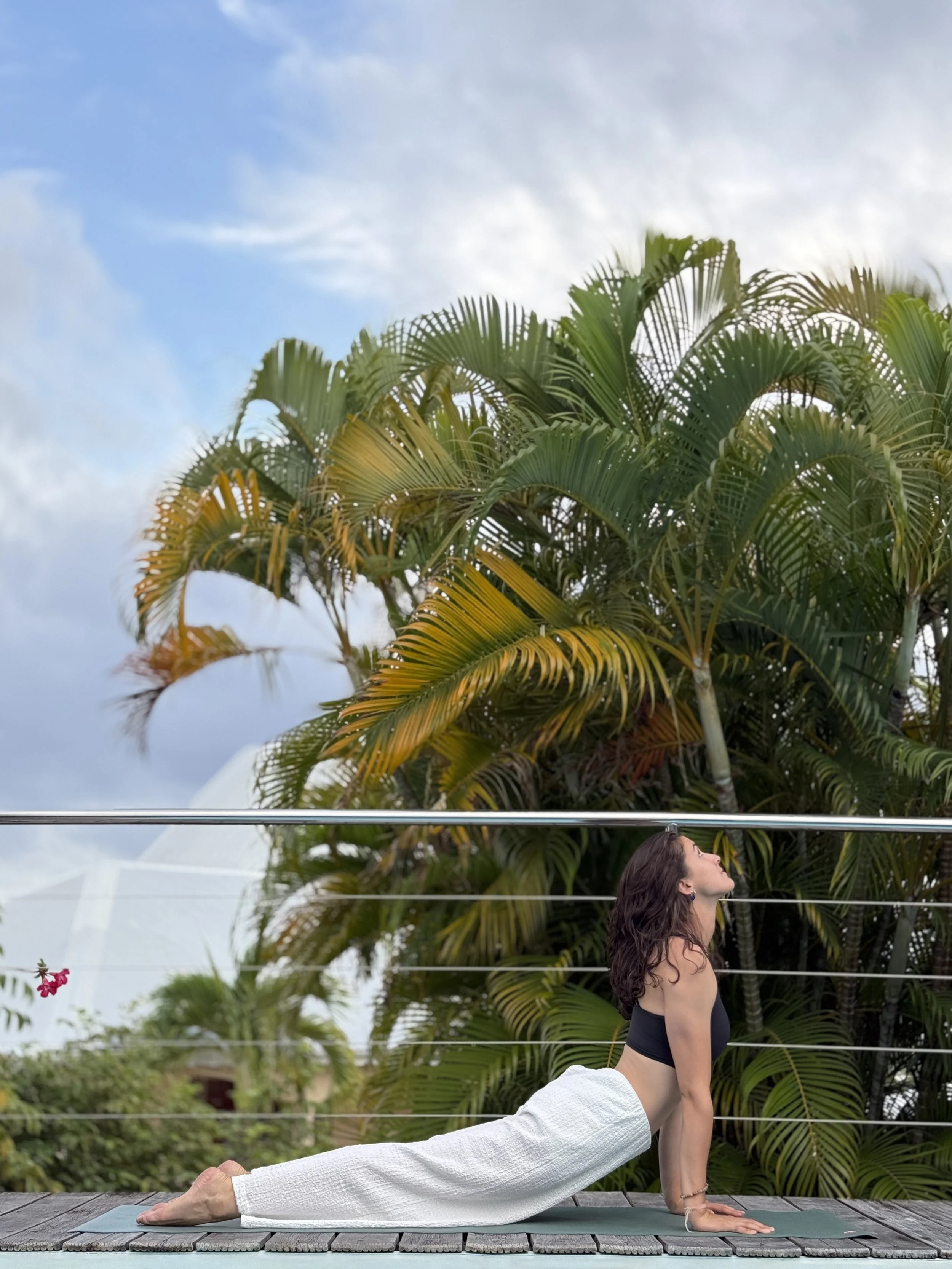 Une femme pratique une posture de yoga sur un tapis près de palmiers, avec un ciel partiellement nuageux en arrière-plan.