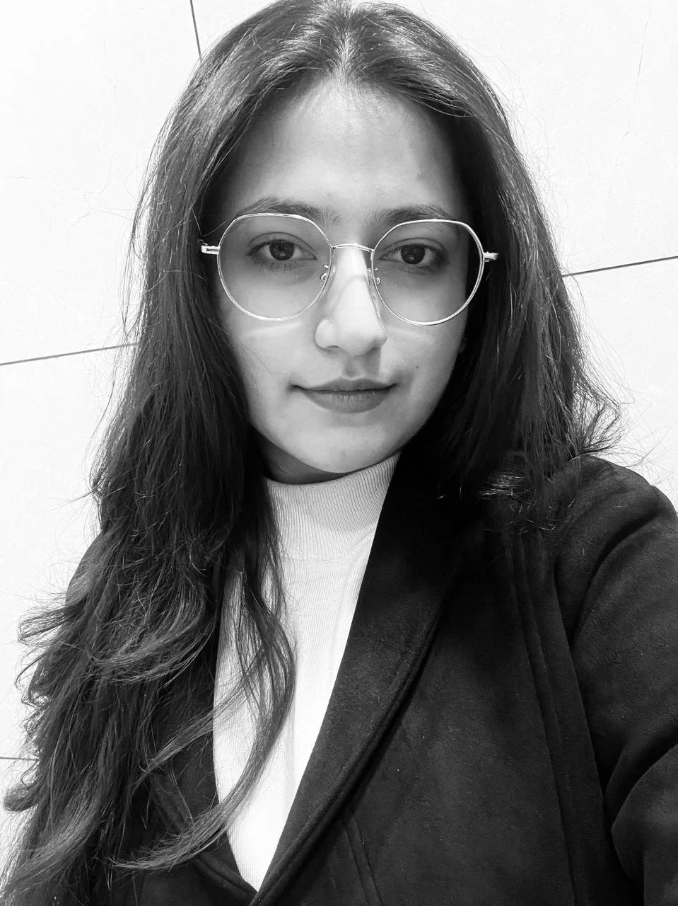 Black and white photo of a young woman with long hair and round glasses, wearing a turtleneck and coat against a plain background.