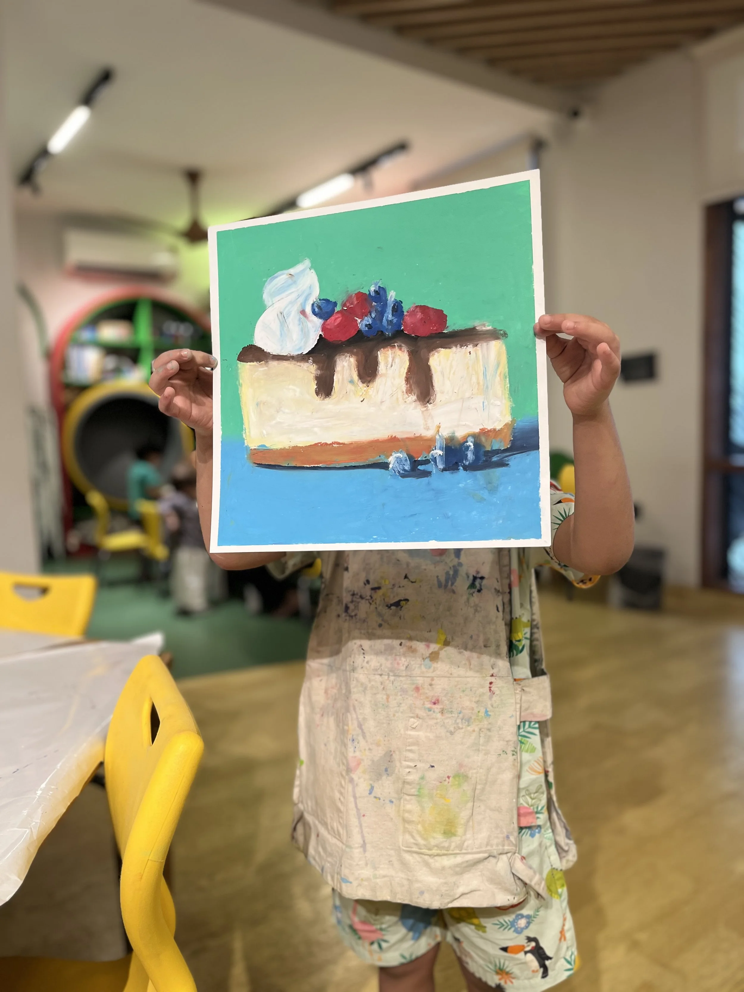 A person holding up a colorful painting of a dessert topped with berries, whipped cream, and chocolate sauce.