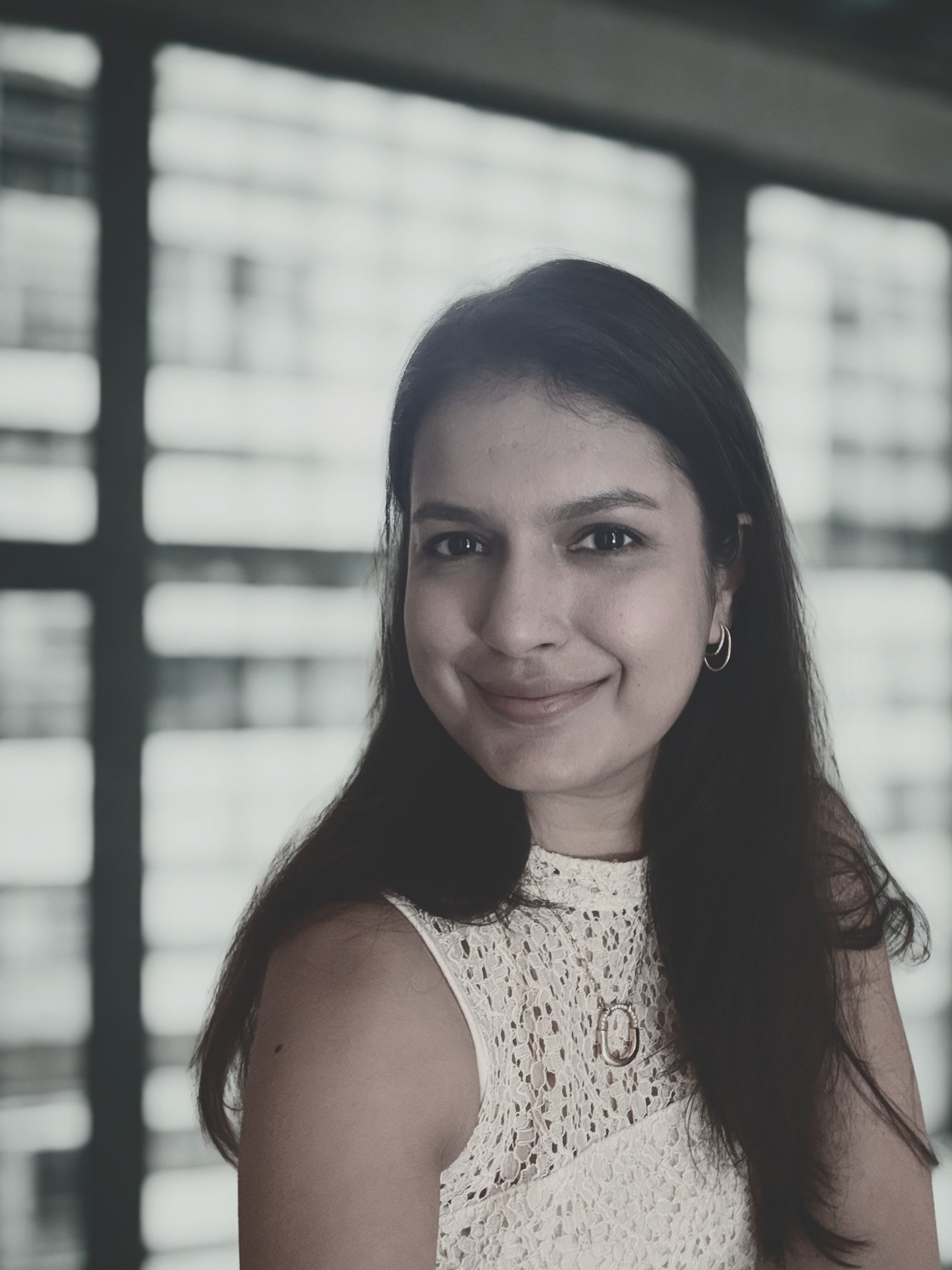 A young woman with dark hair smiling, wearing a white lace sleeveless top, standing in front of a modern building with large windows and horizontal lines.